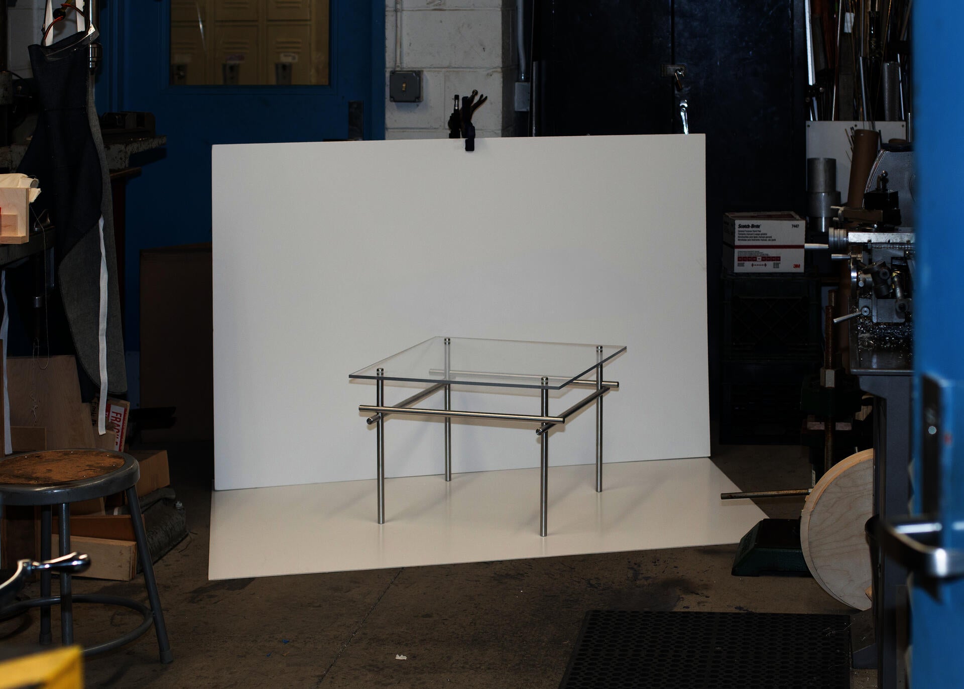A small side table with a clear acrylic top and brushed metal rod frame is displayed against a white backdrop in a machine shop, surrounded by industrial tools, equipment, and workshop materials.