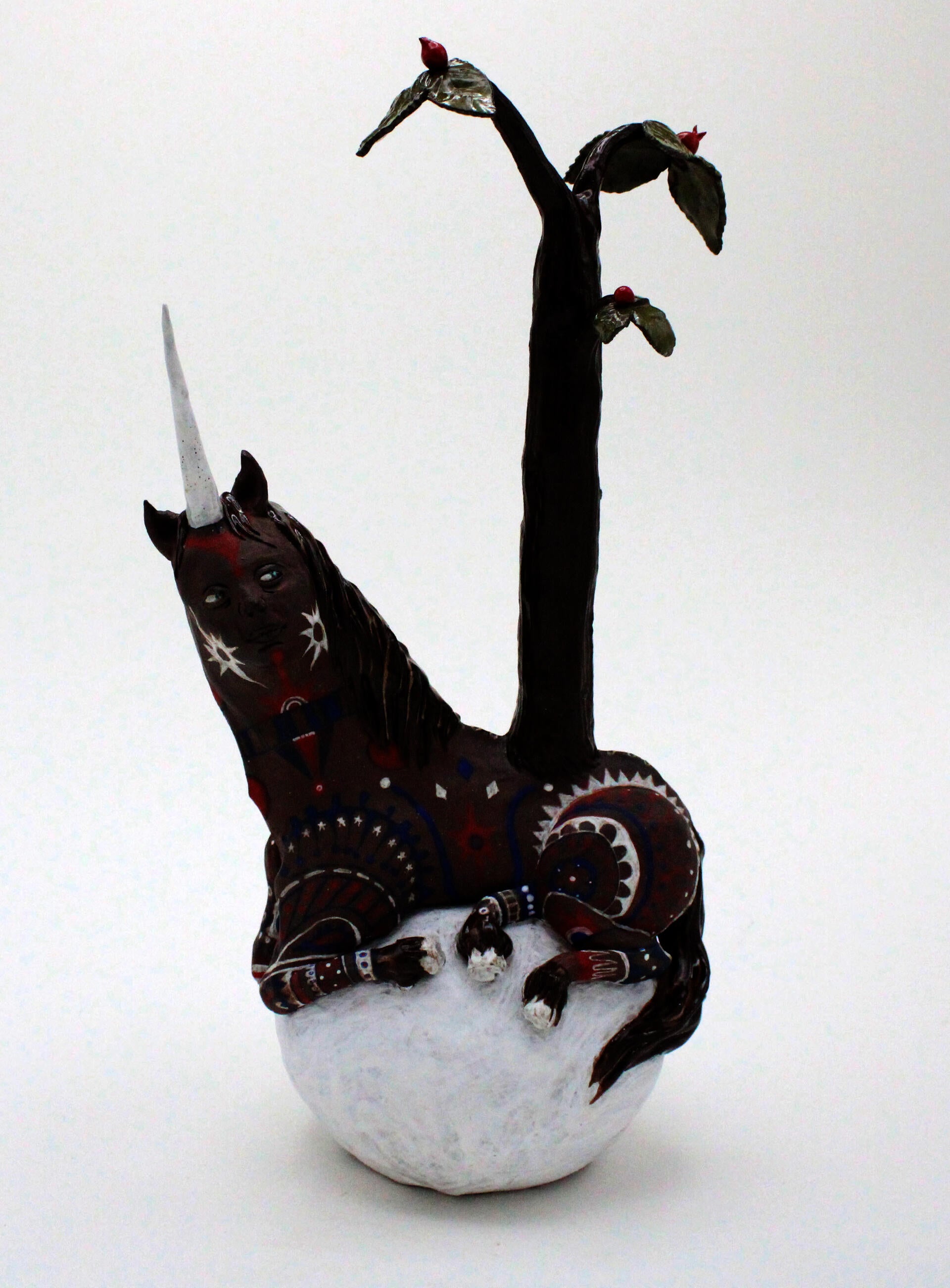 All ceramic: A brown unicorn on top of a white ball. It has the face of a woman and a tree growing through its back. It is covered in patterns inspired by Pictish warrior tattoos.