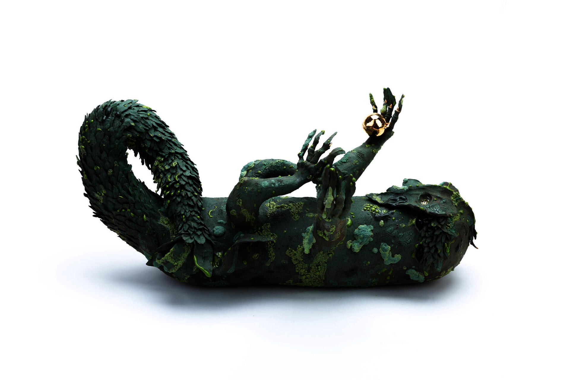 A green four legged creature lying on its back and reaching up to the sky. It has a twisting leafy tail, and is covered in mossy texturing. It holds a golden bell in one hand.