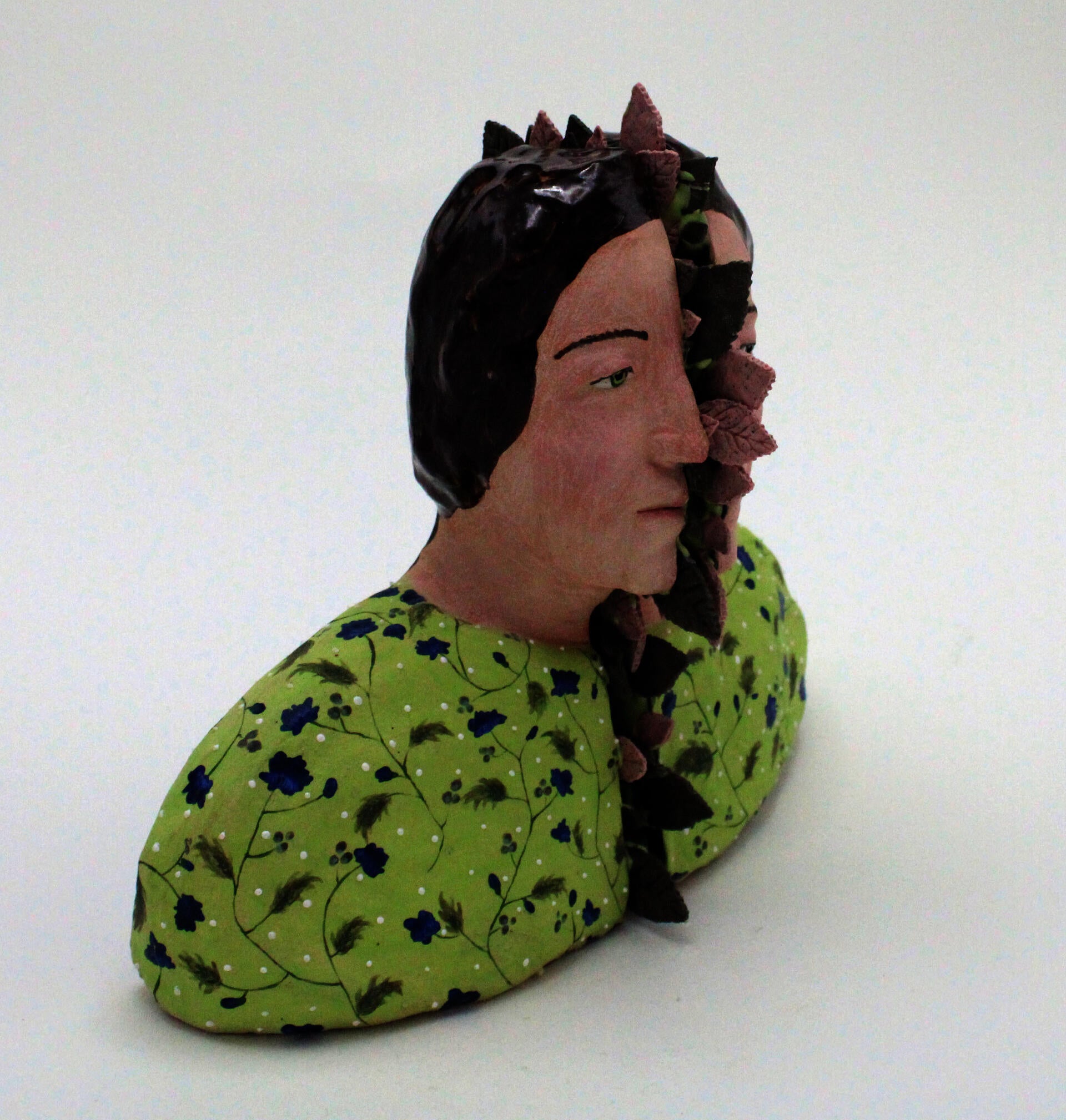 A bust of a woman split in half by green and pink leaves. She is wearing a green shirt with blue floral patterns
