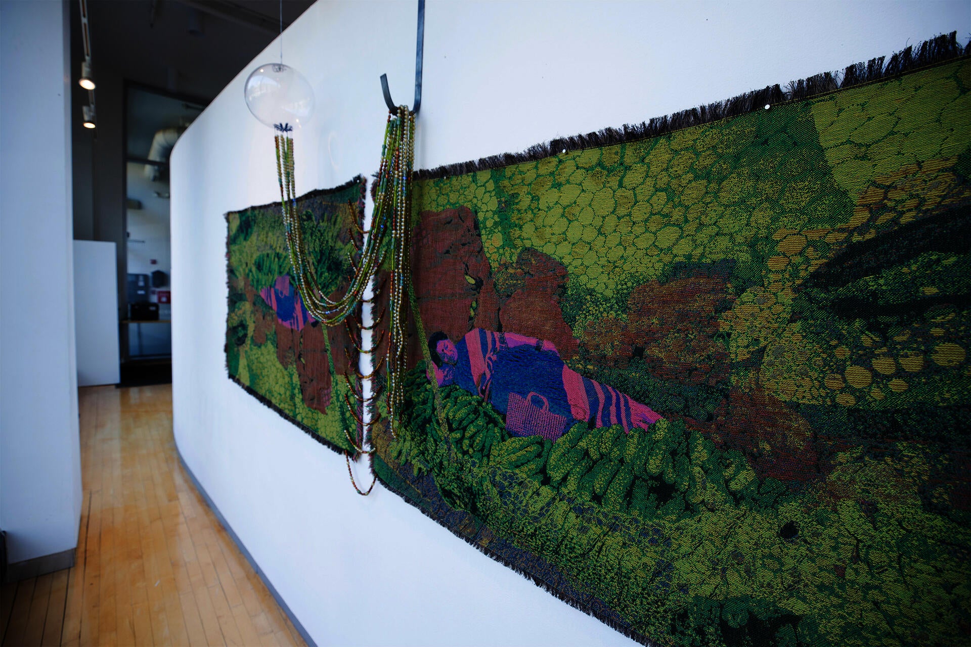 Jacquard-woven textile with lying figure among green forms, with draped glass beads connecting to a hanging glass orb