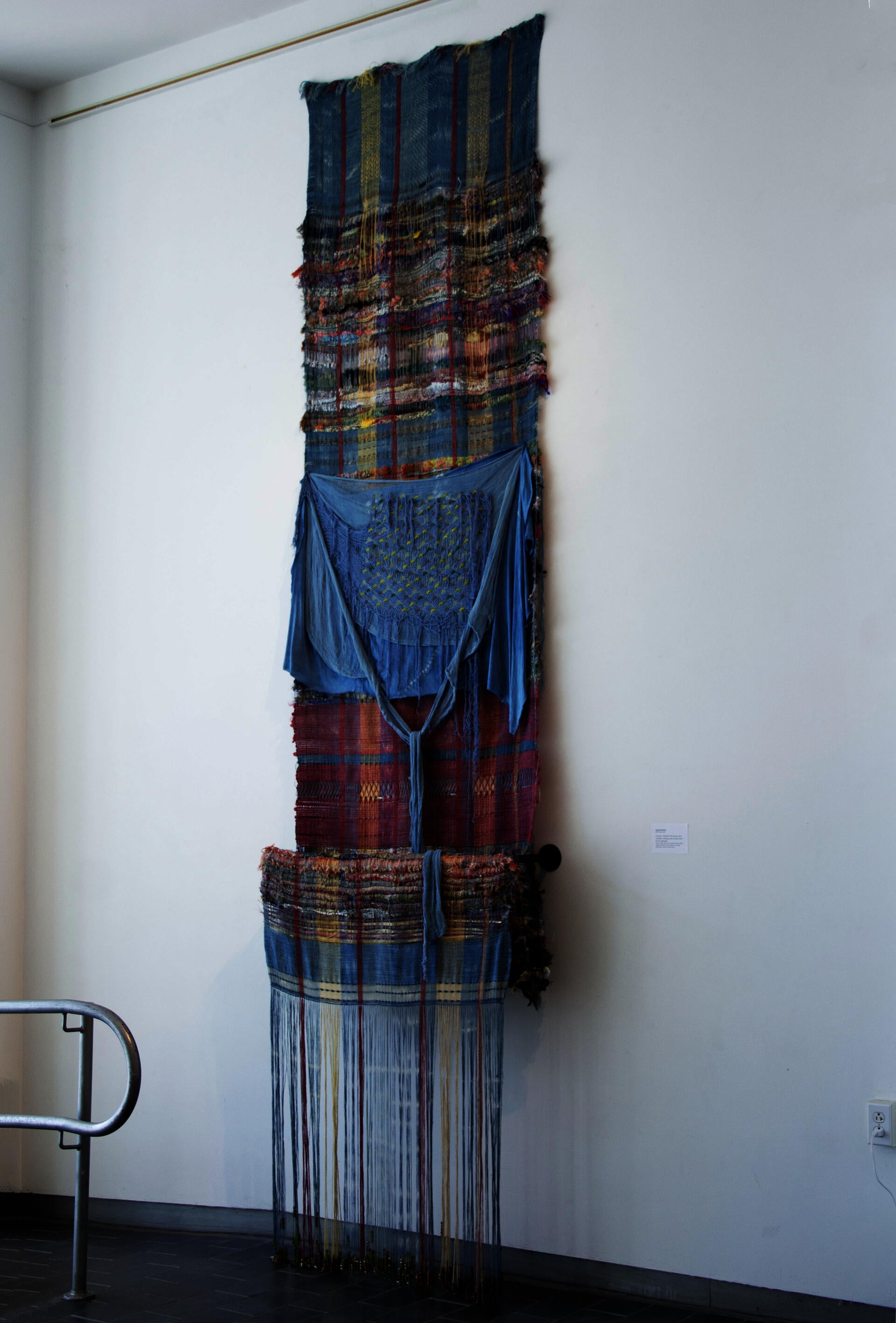 Vertical woven textile installation with layered blue and multicolored cloth, draped fabric, and long hanging thread beaded with glass beads