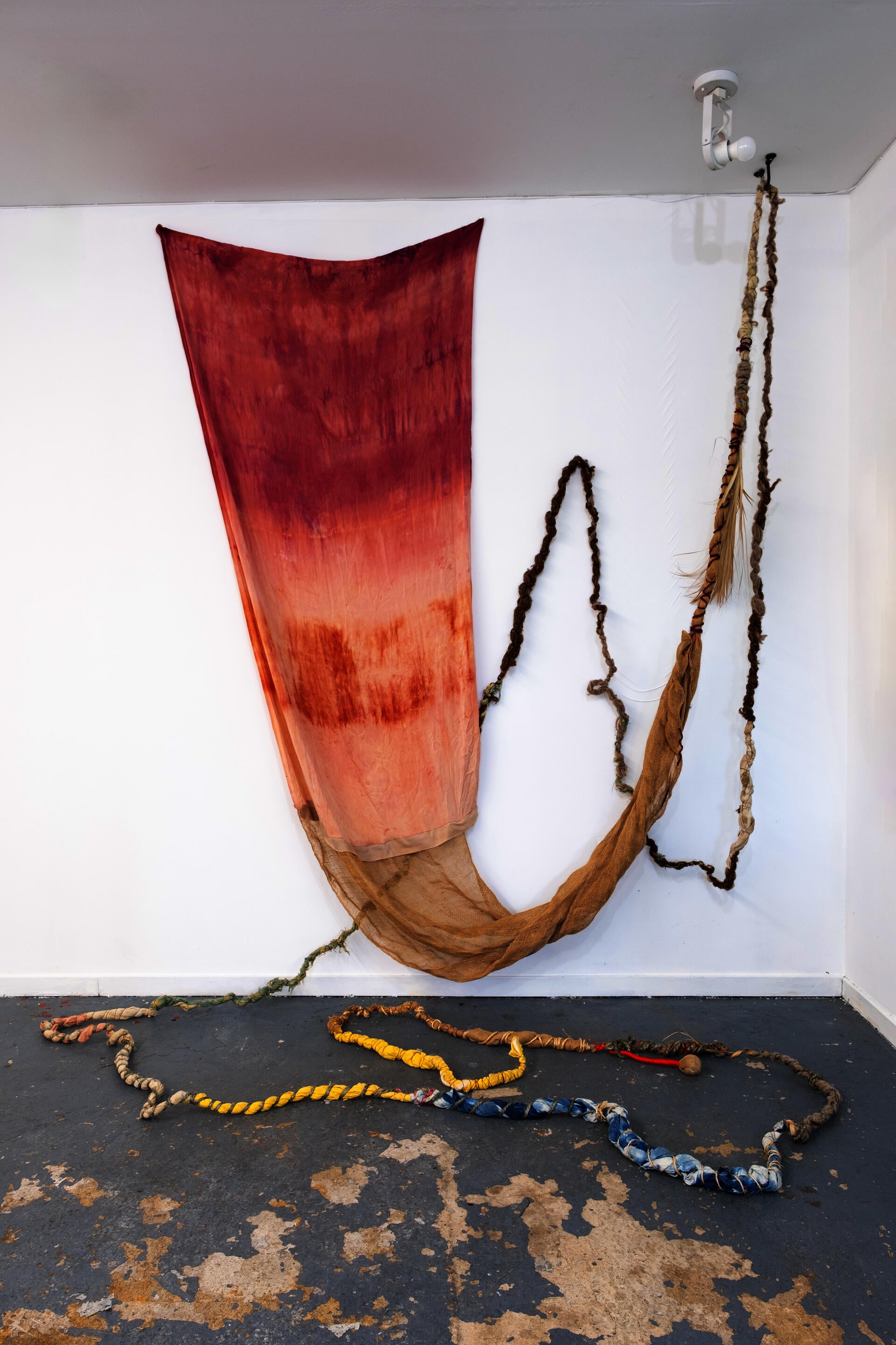 Hand-Natural Dyed textile in red and orange tones draped on wall, with rope-like fibers hanging and extending across the floor with small circle clay forms 