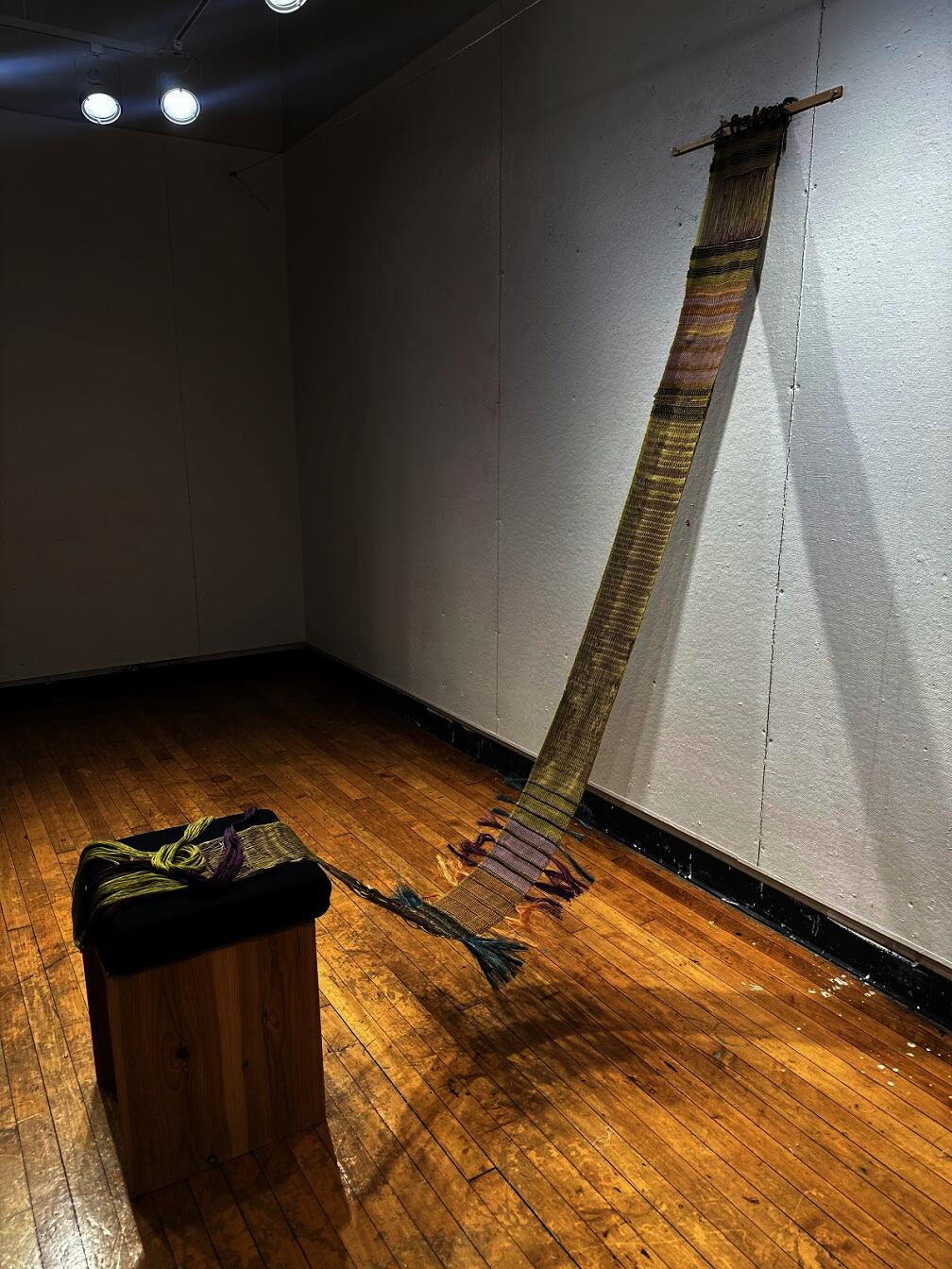 Narrow floor loom woven textile hanging from wall and trailing to floor, with striped patterns, draped onto a wooden pedestal with yarn