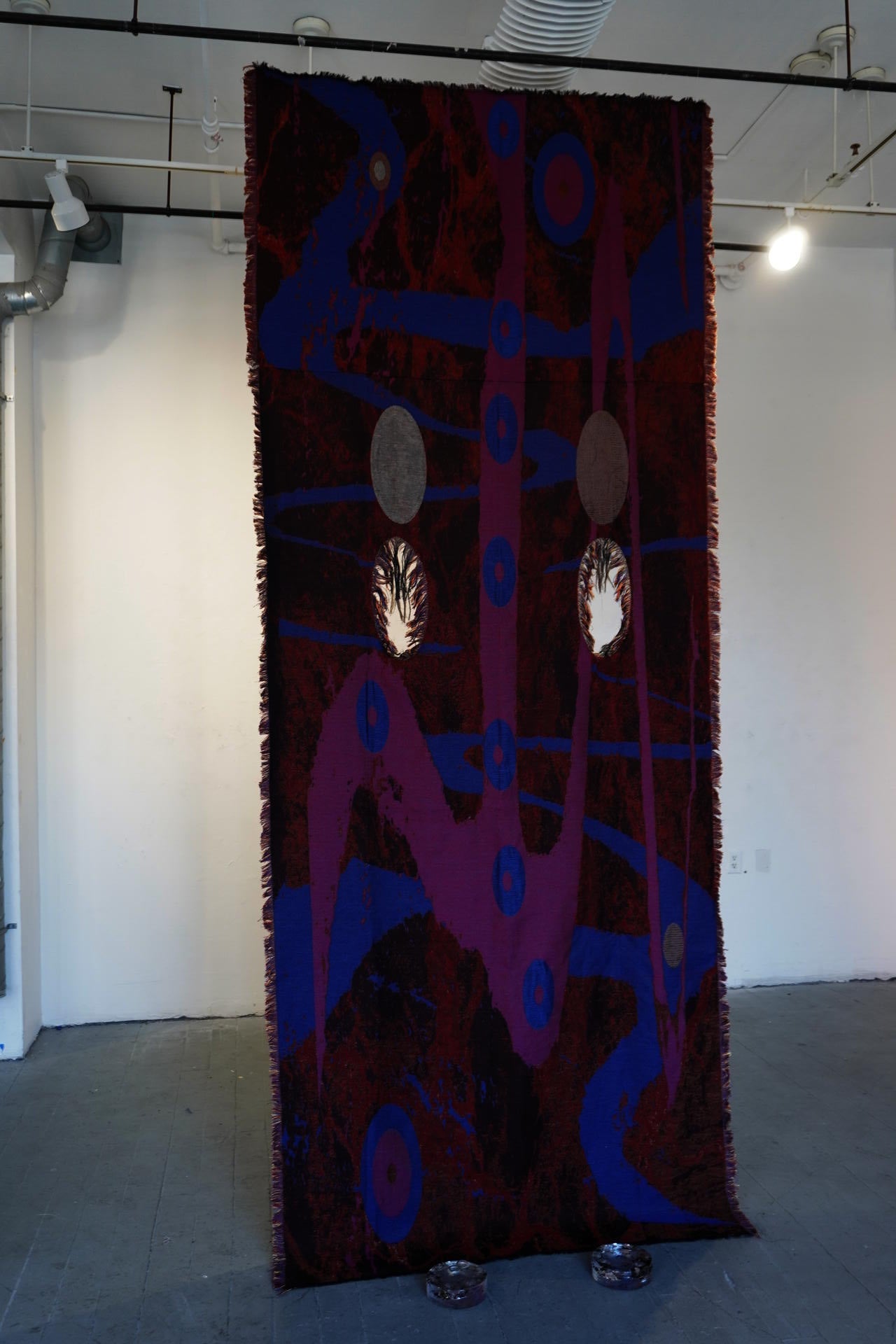 Tall hanging textile with red ground and blue abstract patterns, with reflective oval elements in a gallery space