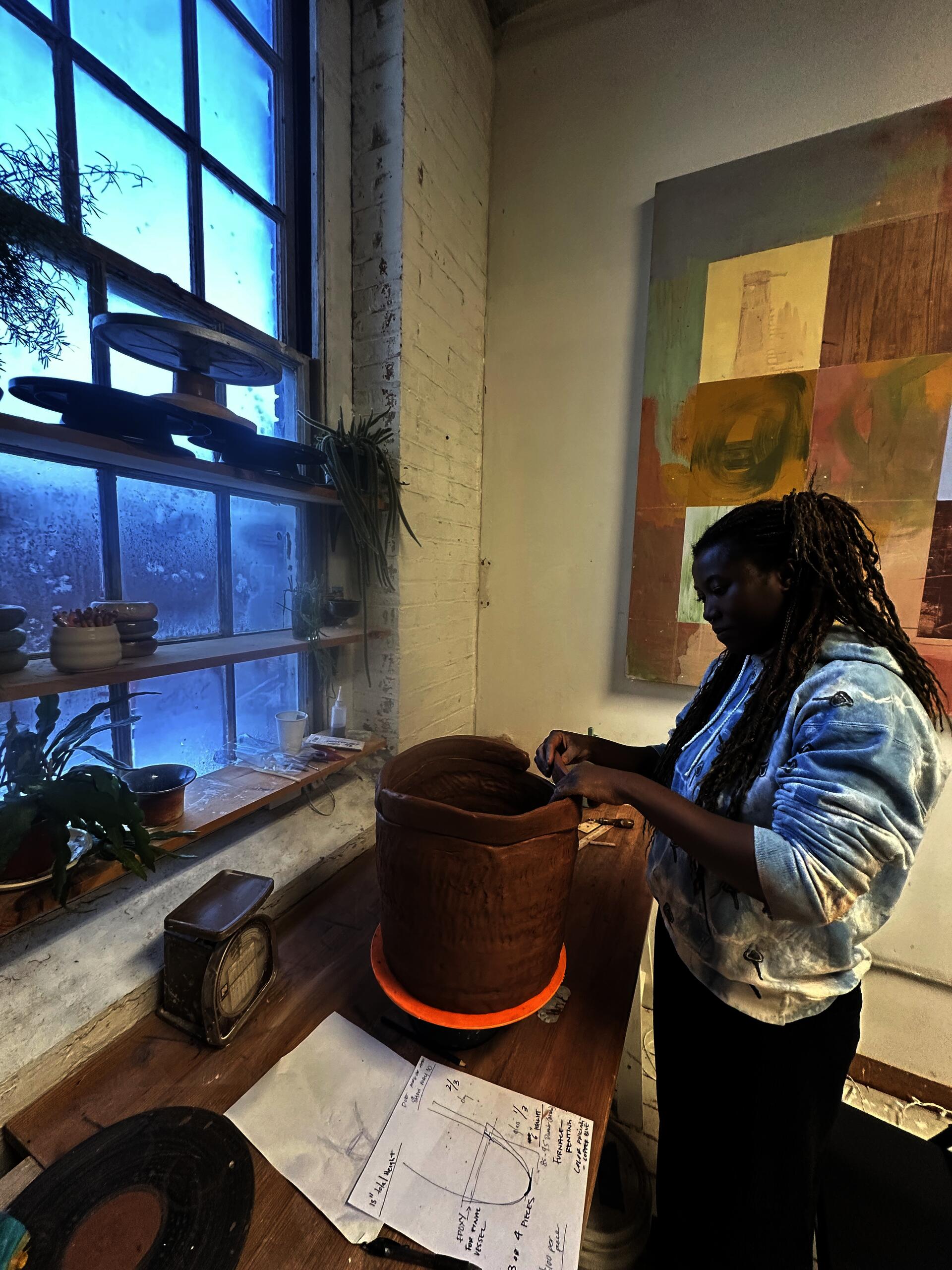 Artist (Asmaa Amadou) working on a large clay vessel at a studio table by a window, with tools, plants, and sketches nearby