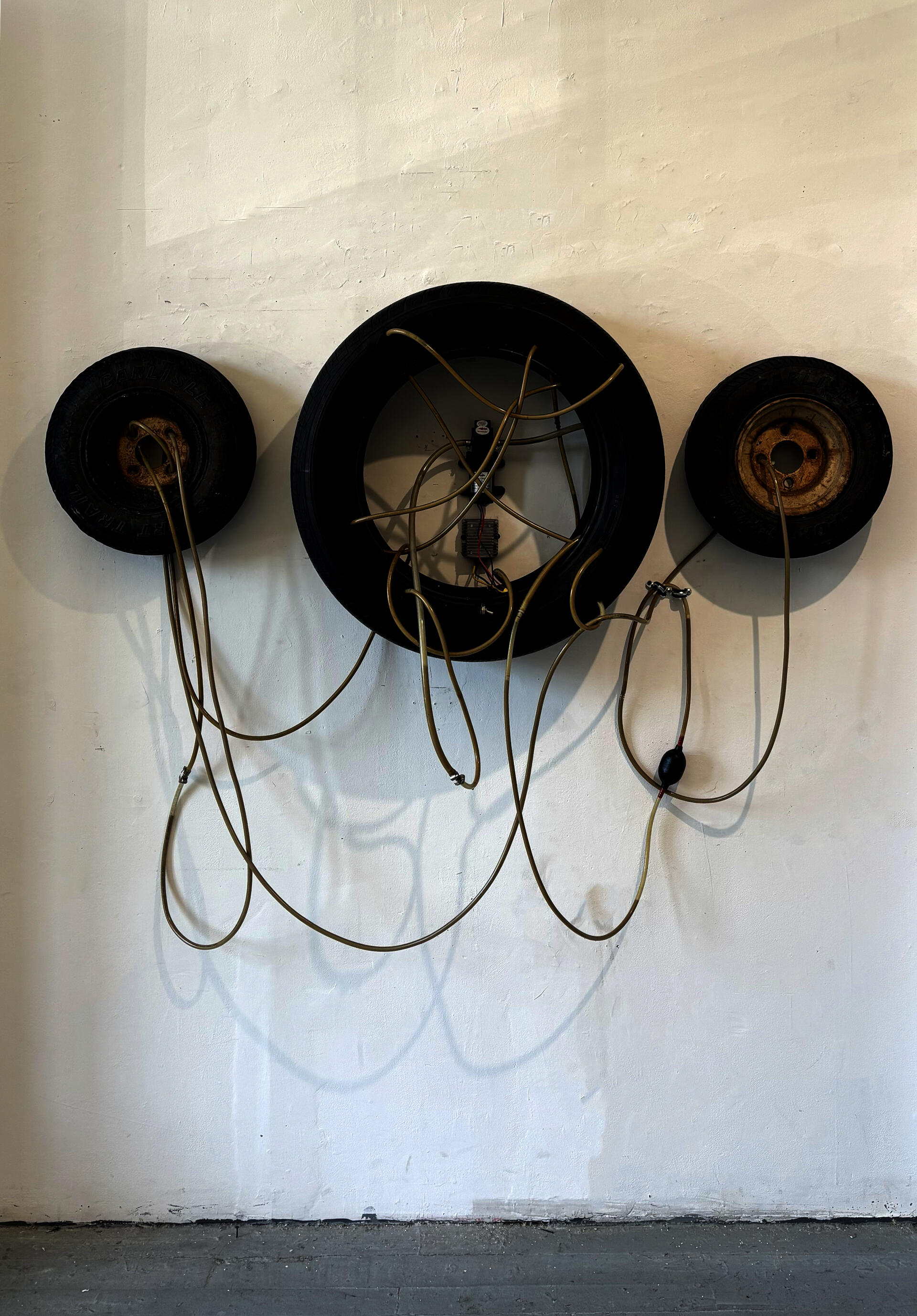 Three tires are assembled in a triptych format and are connected by a series of tubes and siphons. A motor is placed in the middle of the system and pumps liquid through allowing for some to leak out. 