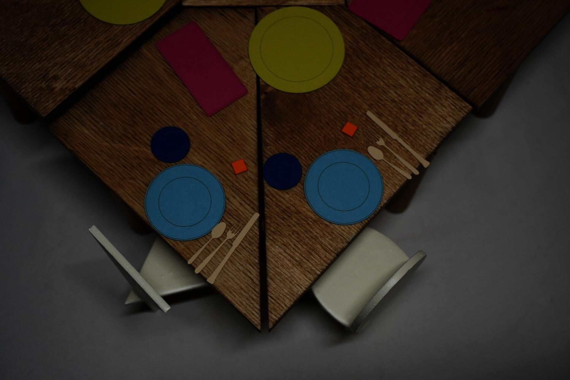 A corner of a table is shown from overhead view, where the pieces are broken in two, and  two different chairs sit adjacent to each other. Colorful kitchenware is scattered across the table surface. 