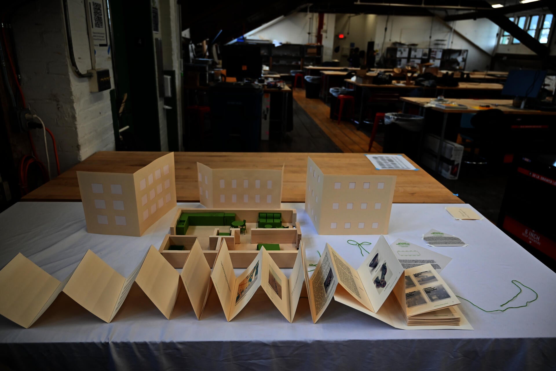 1/2" scale wooden model of one bedroom apartment and green paper furniture. Walls at 1/2 height. Apartment sits in front of paper building elevations. Accordion book spans the studio desk in front.
