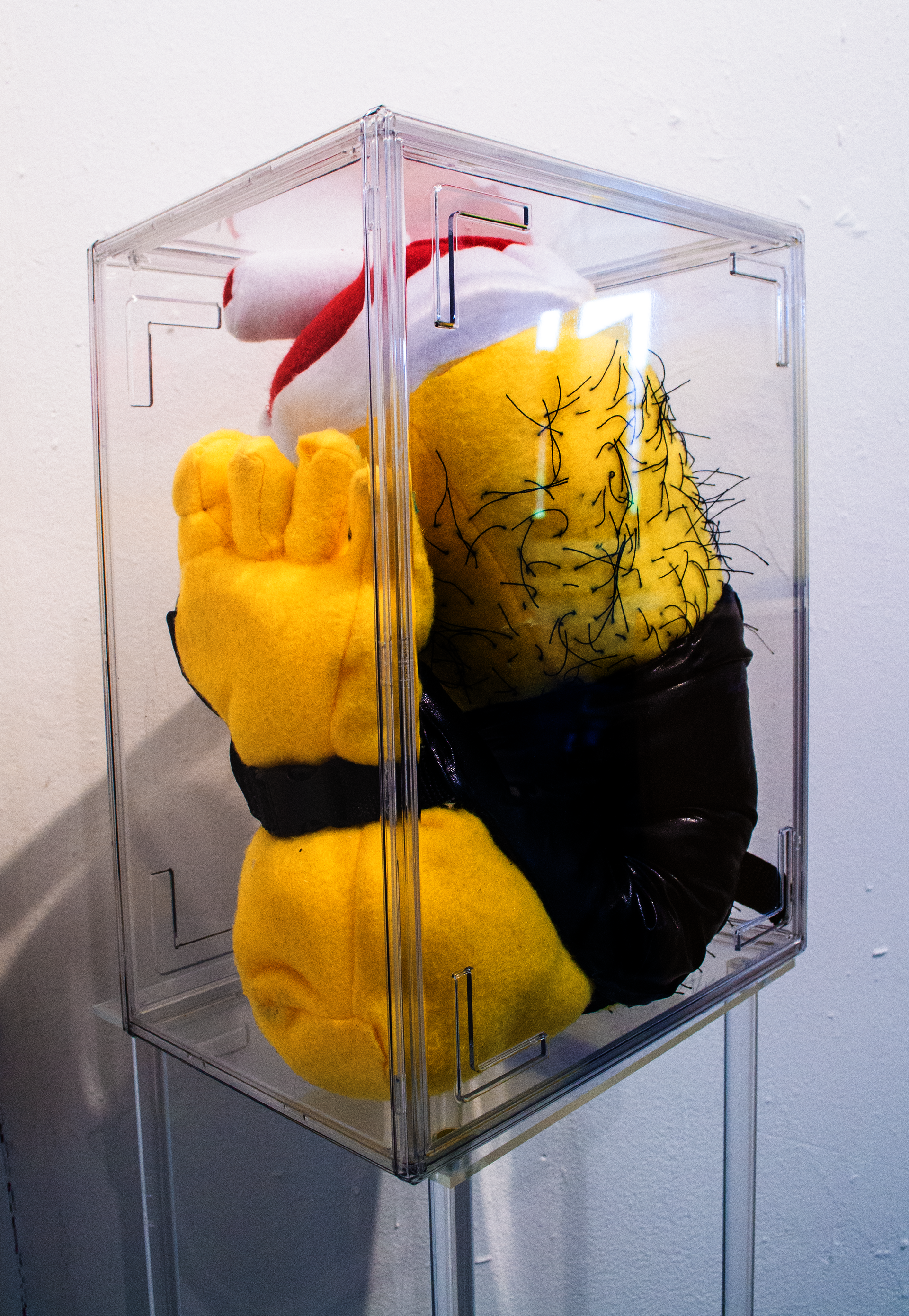 A clear acrylic case housing a plush, bright yellow severed foot. It is wearing a gladiator's boot. 