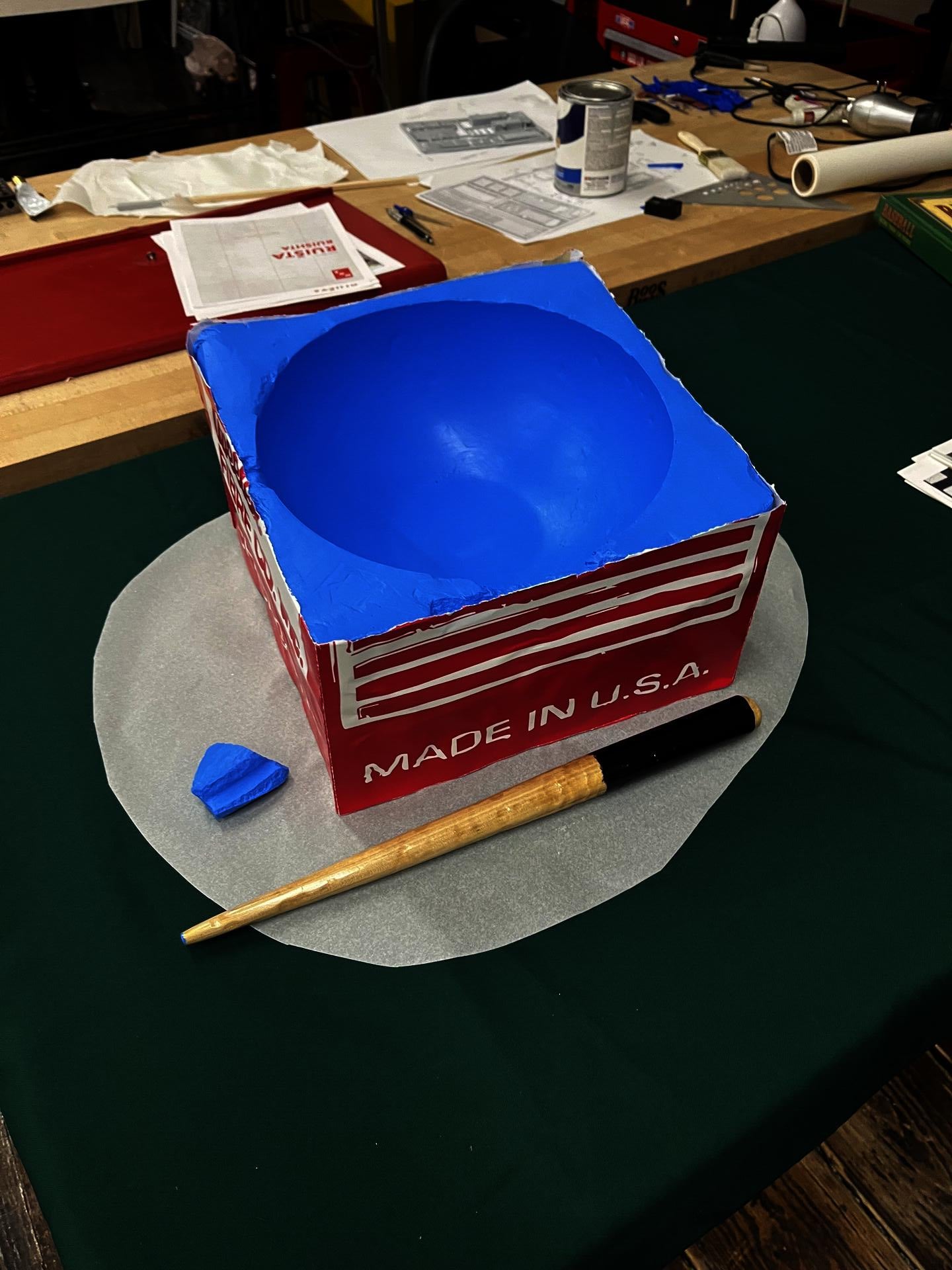 Process photo of an oversized blue pool cue chalk beside a miniature pool cue on a worktable. 