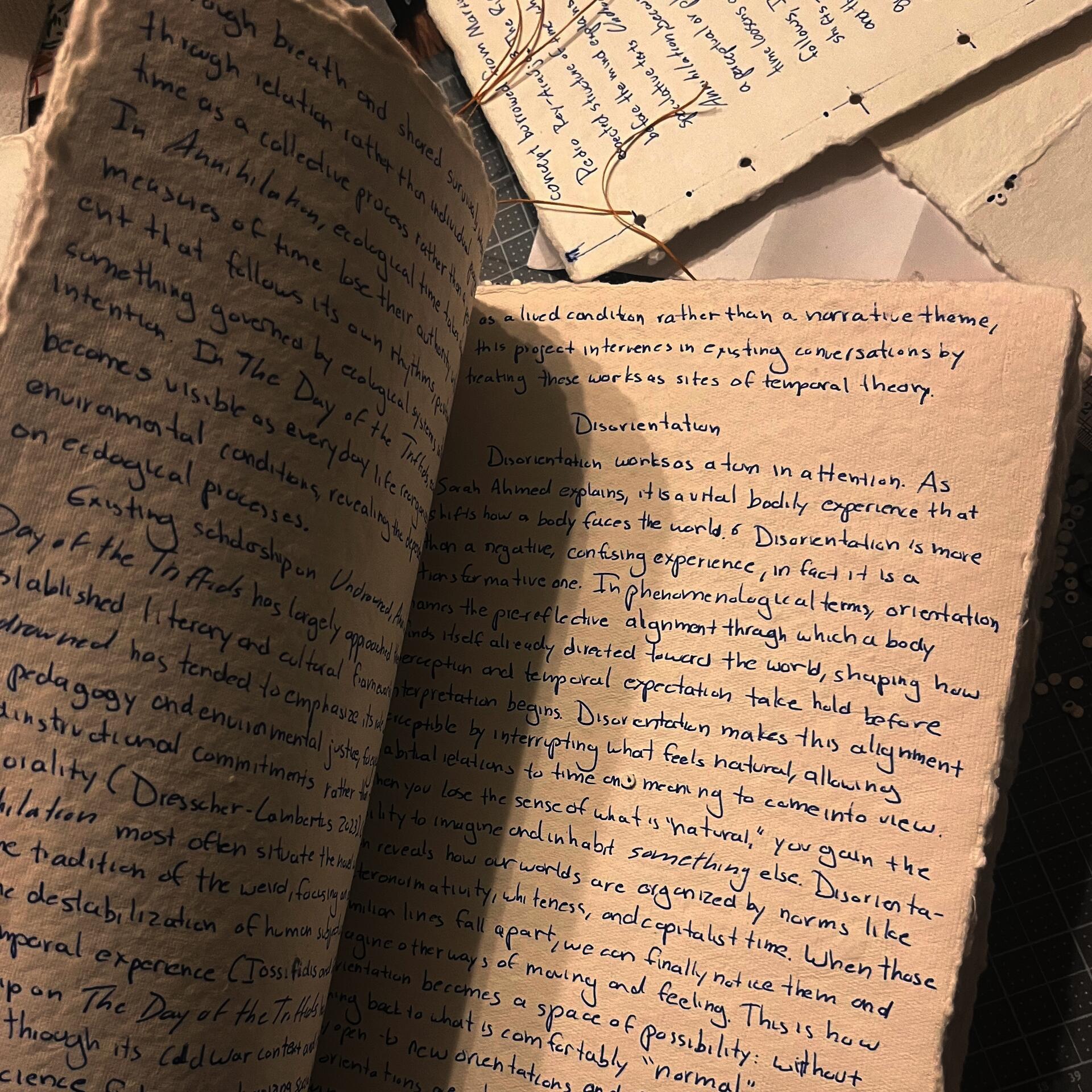 Open rough, cream paper thesis manuscript handwritten in blue ink sits on a cutting mat beside stitched pages and loose thread. Visible section titled “Disorientation” discusses Sarah Ahmed, phenomenology, and queer temporality.