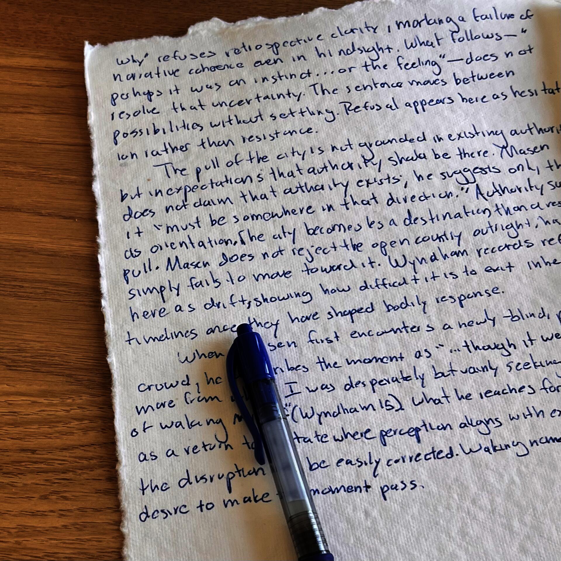 Deckled cream paper covered in blue handwritten thesis prose rests on a wooden table beside a blue pen. Text discusses refusal and uncertainty in relation to temporality and perception