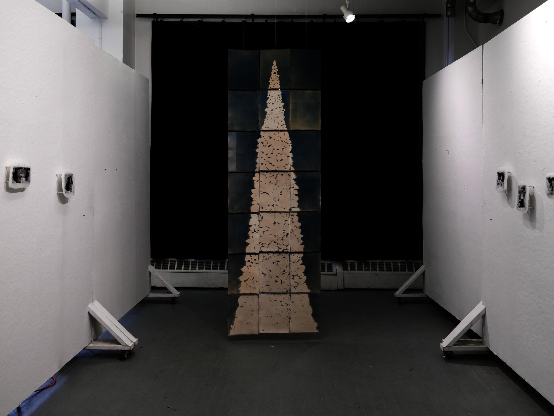 Installation view with a tall cyanotype stone tower hanging between white walls, with small paraffin photo works on both sides.