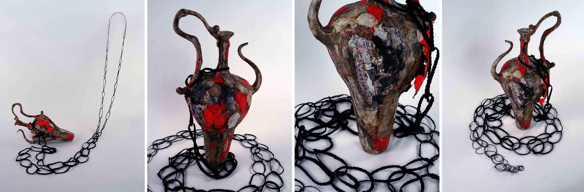 A large paper vessel with layered and burned edges sits low on the ground, surrounded by necklaces made from strands of dark hair. The vessel rises upward in sharp folded forms resembling flames made of burnt paper.