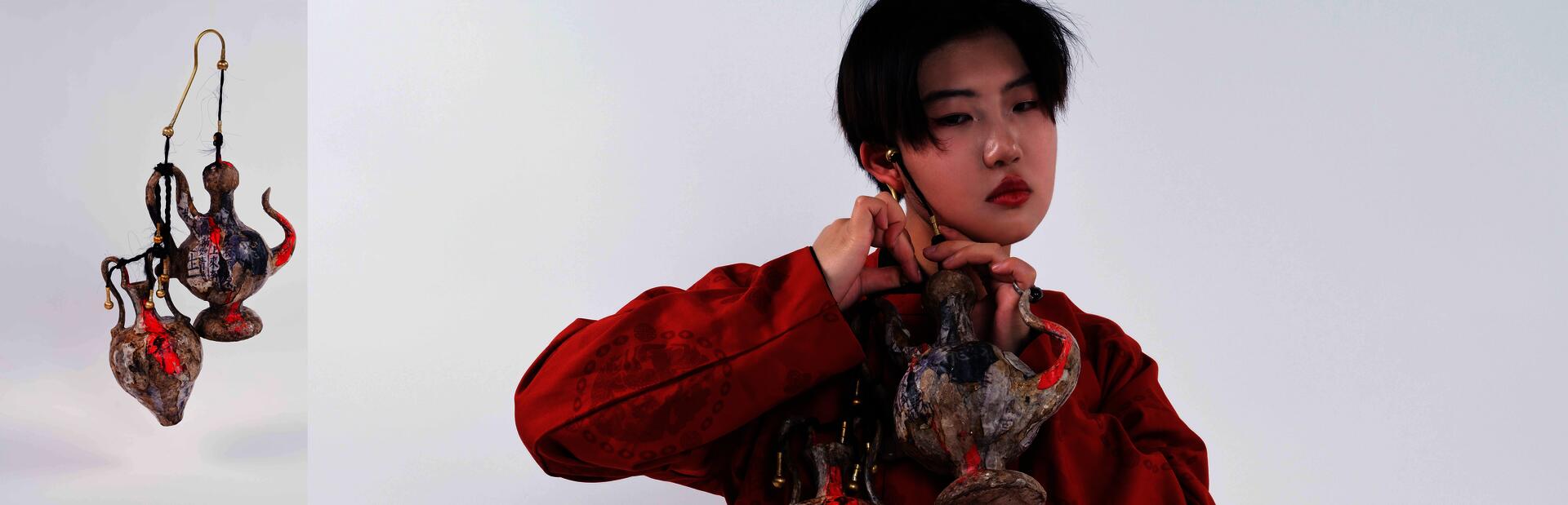 A model wearing a red traditional Chinese Tang dynasty round-collar robe demonstrates how to wear two oversized ear-hanger jewelry pieces. The model hangs the large ear ornaments from the ears and stands in a static pose.