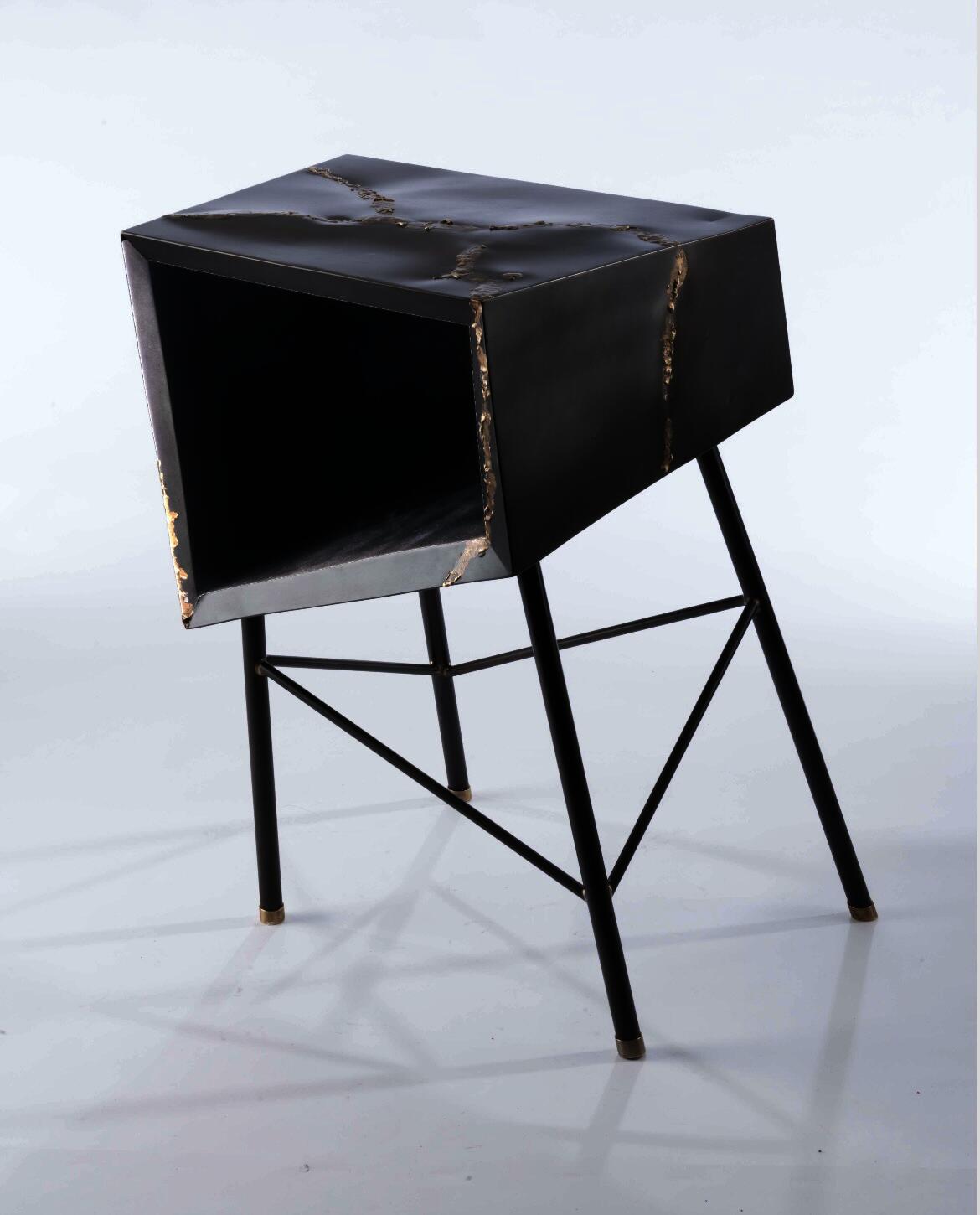 dark steel angular box sitting atop thin metal tubes arrange that seem askew, each connection has a gold fillet, there are brass feet and the corner shows melted bronze dripping down from the top surface