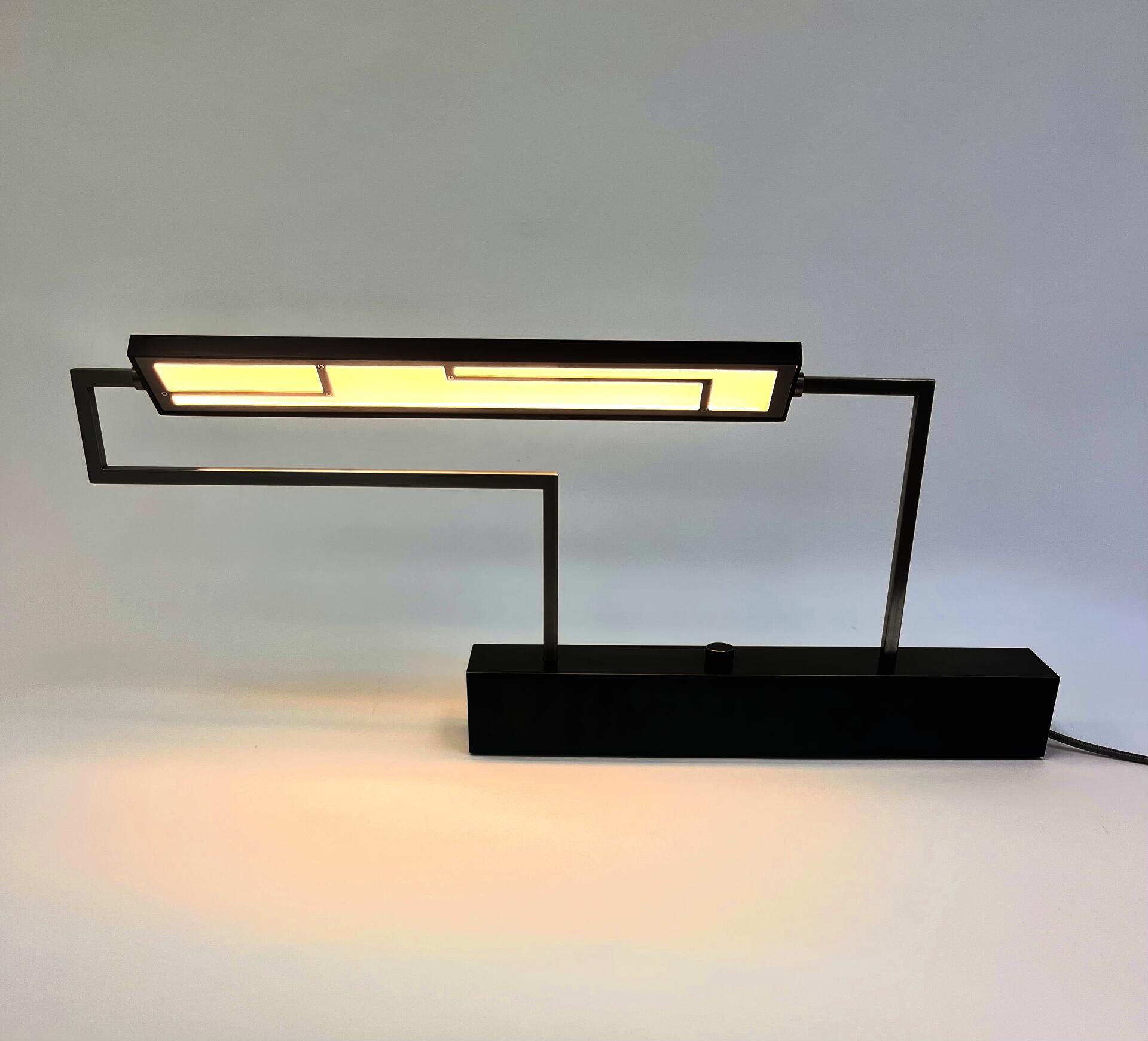 dark blue rectangular light with a patterned metal cover over diffuser, thin polished steel tube supports, larger dark blue rectangular base with a round steel knob for dimmer switch all corners highlighted by polished steel 