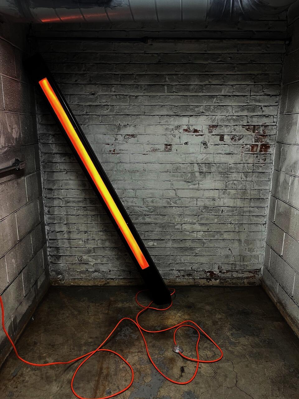 metal beam placed at very precarious angle in corner there is an orange beam of light on the front facing side of the large steel beam, and a jumble of extension cord at the base