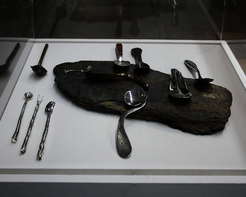 a series of spoons made from a variety of metals placed on and around a flat rock all arranged on a white pedestal enclosed in acrylic