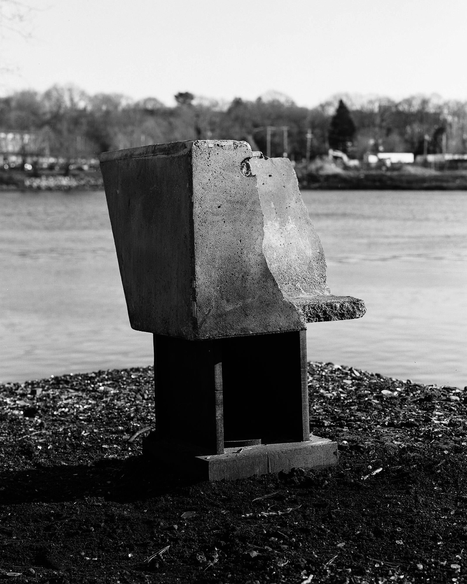 black and white image of concrete sitting atop rusty I-beam metal piece located on the bank of a river