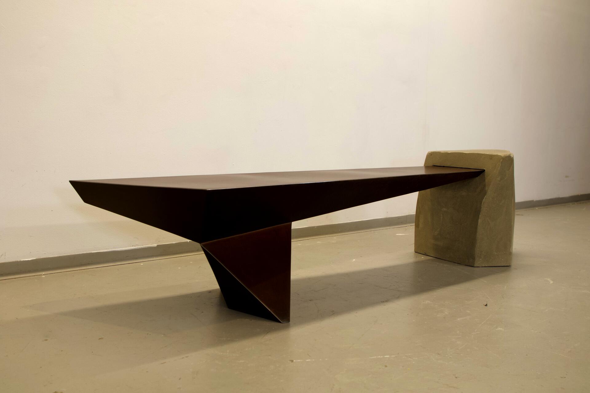 Angular rusty metal bench attached to angular concrete mass, corners of rusty metal have been selectively polished to highlight the edges