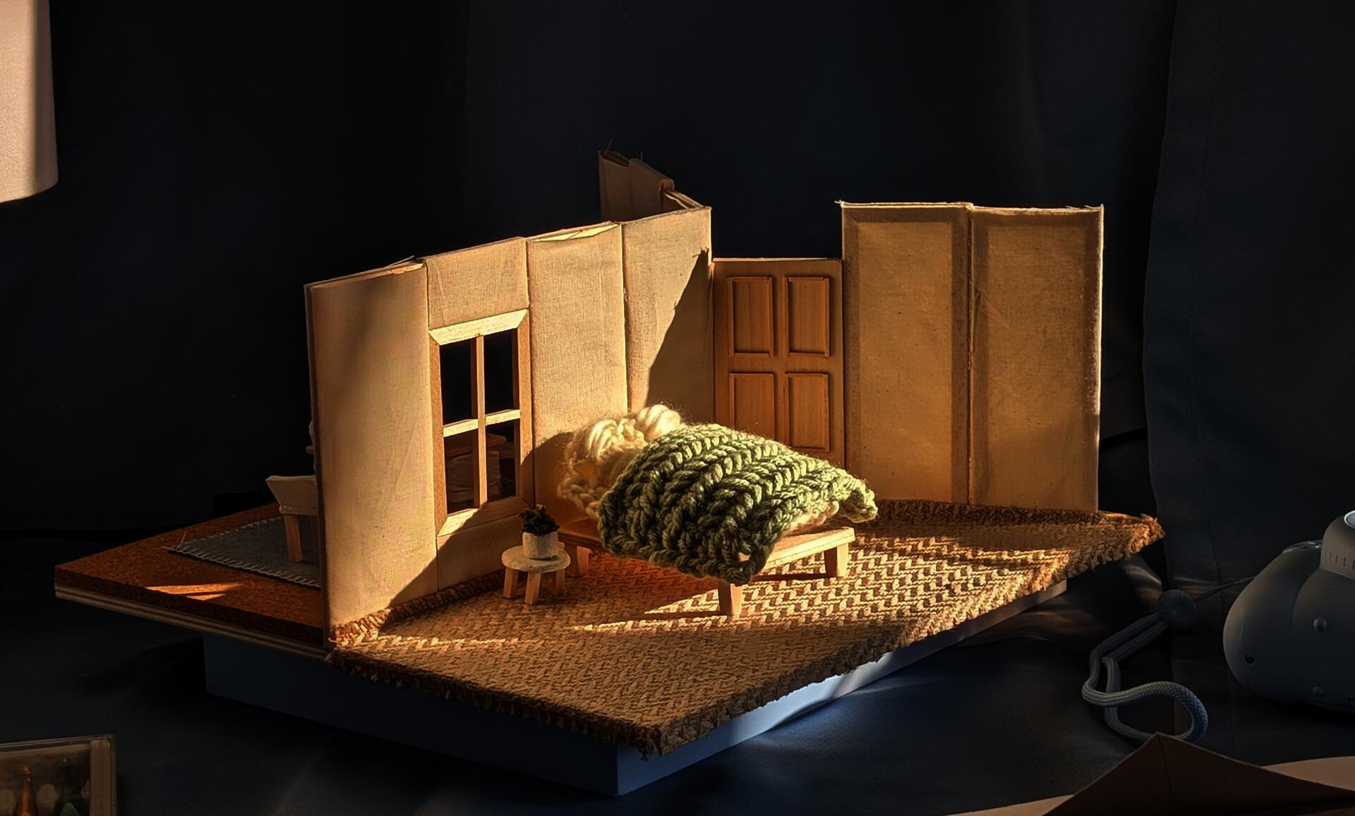 A three way stage model made of textiles, hand knits and crochet. It depicts a bedroom, kitchen, and front porch.
