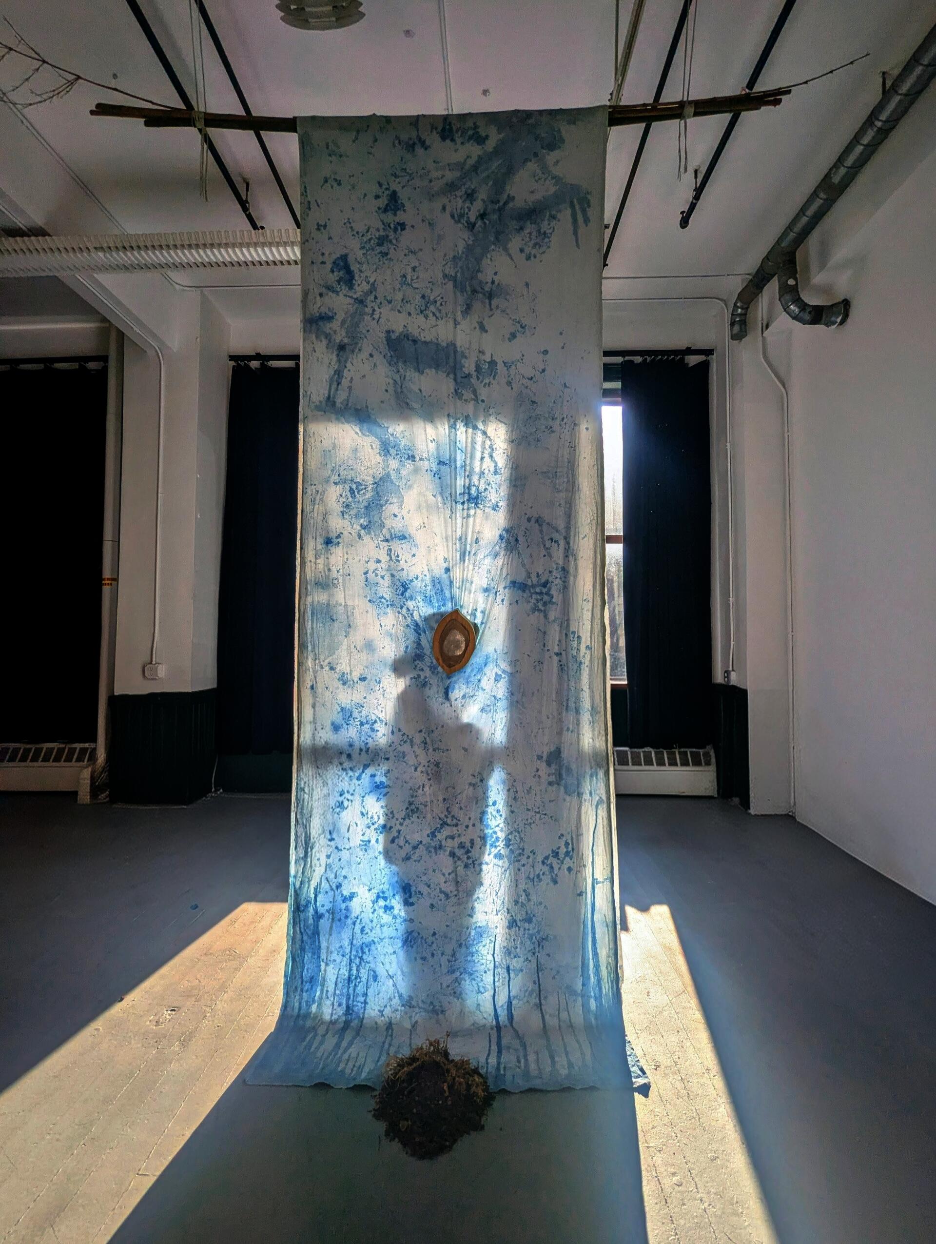 Floor-to-ceiling white cotton draped like a runner, marked with indigo body prints. Backlit to reveal a person's shadow silhouette with a heart-shaped glass above at the center, with dried soil and plants beneath.