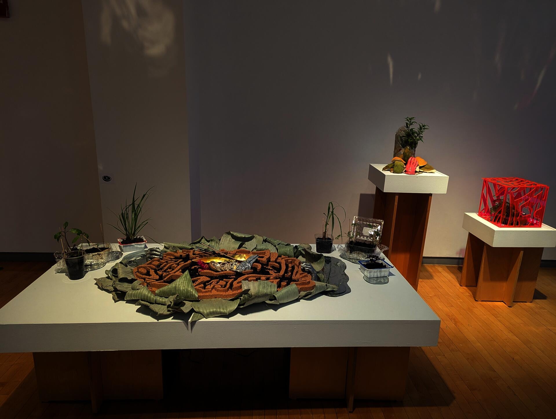 Batik-patterned altar covered in black rice and protective talismans, flanked by live herbs and banana leaves. Two separate pedestals display a woven vessel and a neon-lit batik cube with plants. 