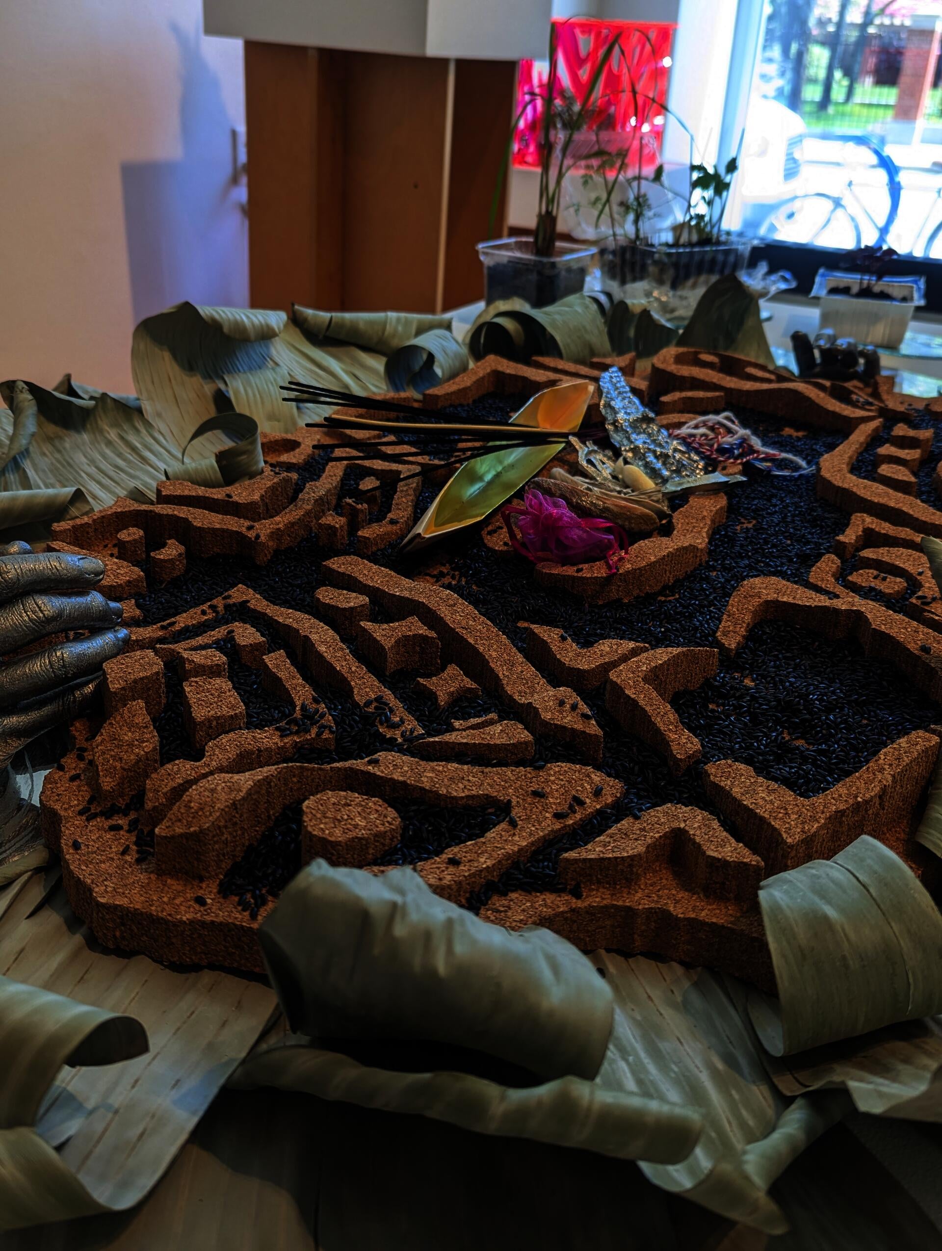 Angled side detail view of Batik-patterned altar covered in black rice and protective talismans, flanked by live herbs and banana leaves.
