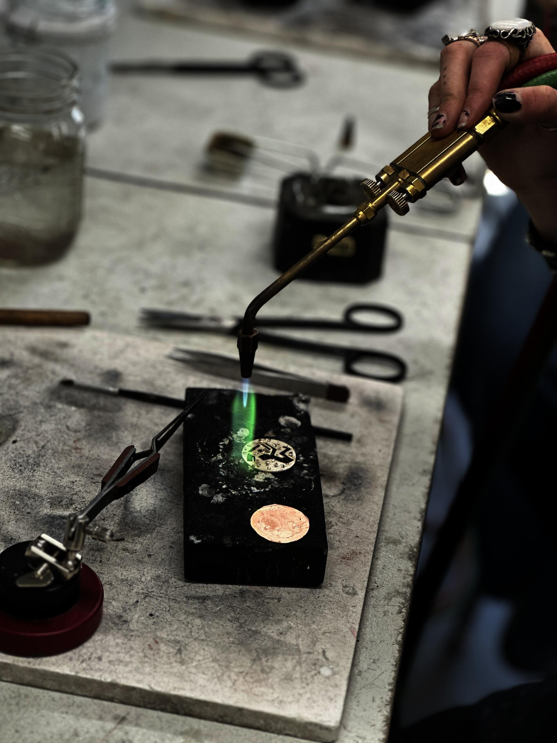 A hand holds a thin torch with a green flame over two copper circles.