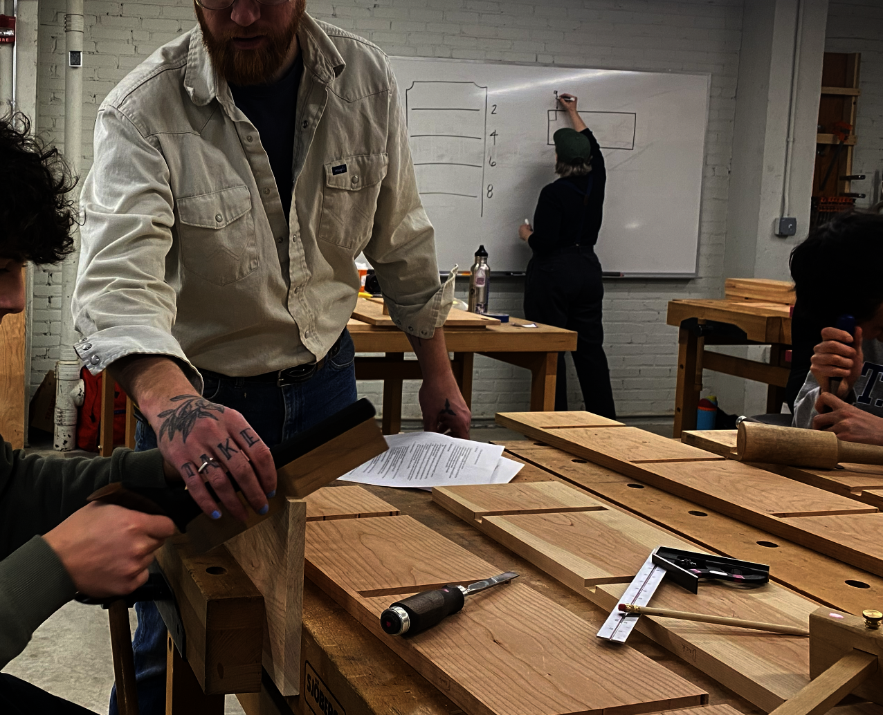 A bearded man holds the saw blade for a teenage boy cutting through a plank of wood in a woodworking classroom.