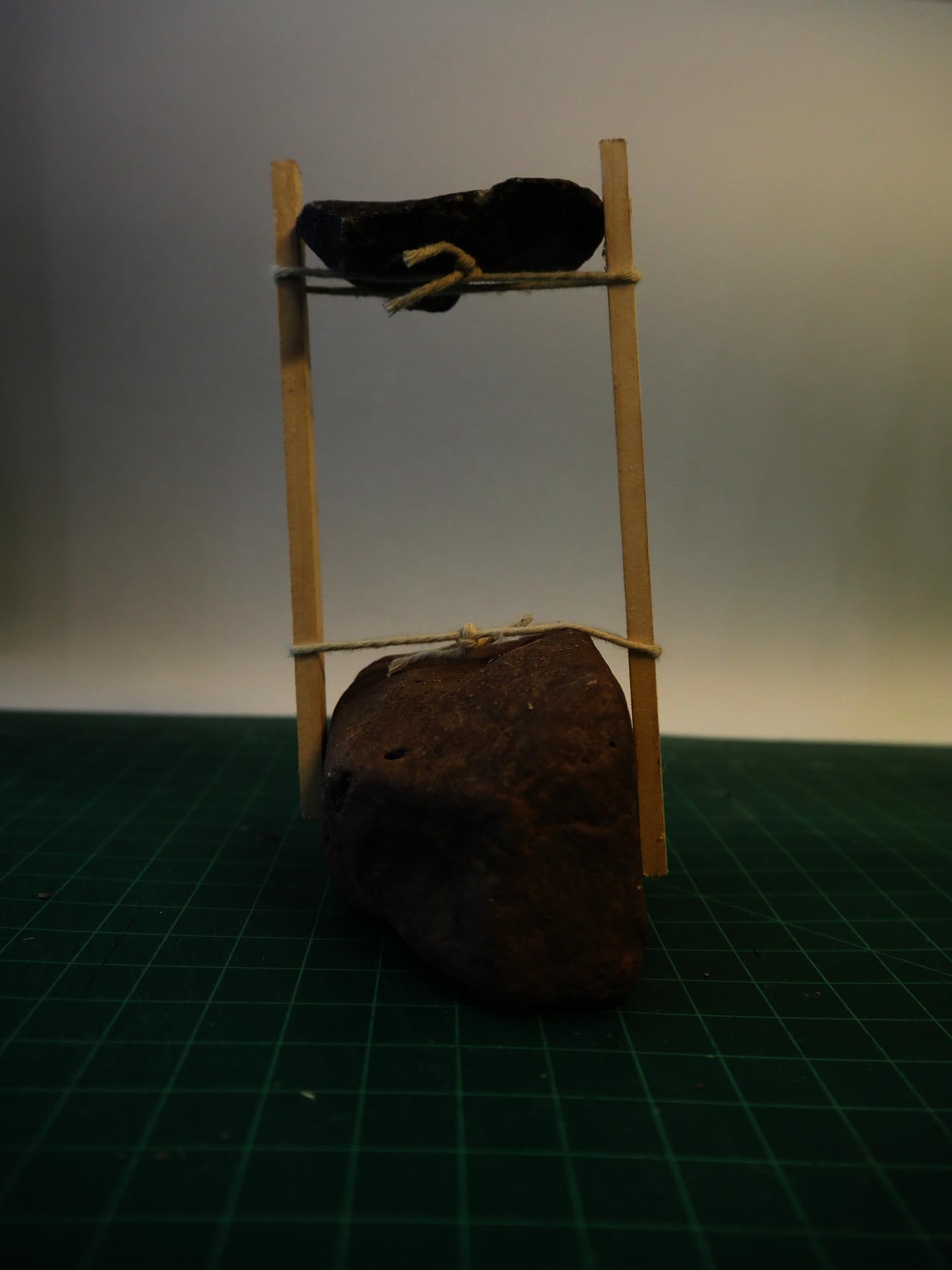 Two wood sticks and string support an elevated rock, anchored by rock on the ground.