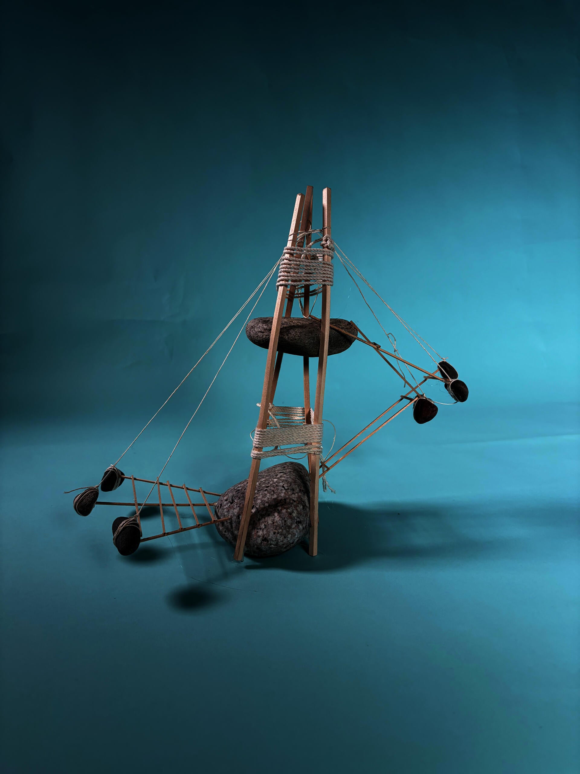 A model made of rock, wood and string. A larger rock on the ground and a smaller rock in the air. Other structures emerge radially, hanging from the top: a deck for research spaces held up by hanging rocks & a set of stairs leading to the top rock platform.