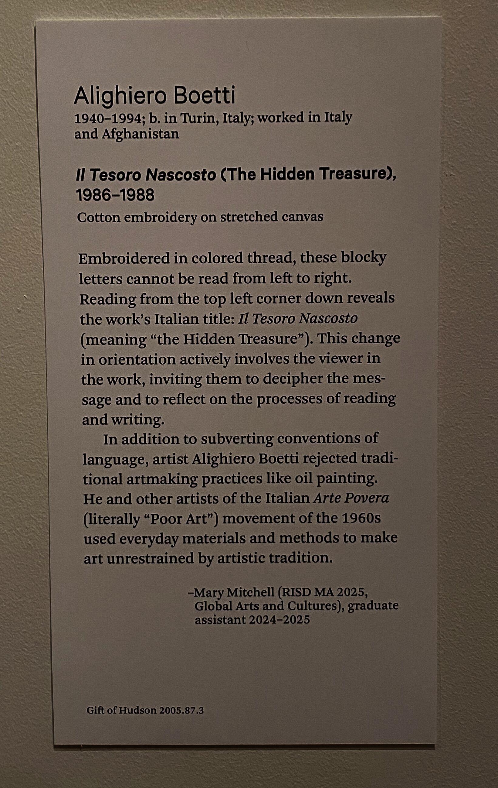 A label affixed to a white wall with two short paragraphs featuring biographical information about the artist, the title of the work, and a list of materials used. Below are two paragraphs of interpretive description that explain the viewer's involvement through reading the work's message and details about Boetti's practice. A signature credits a RISD grad student as the author. 