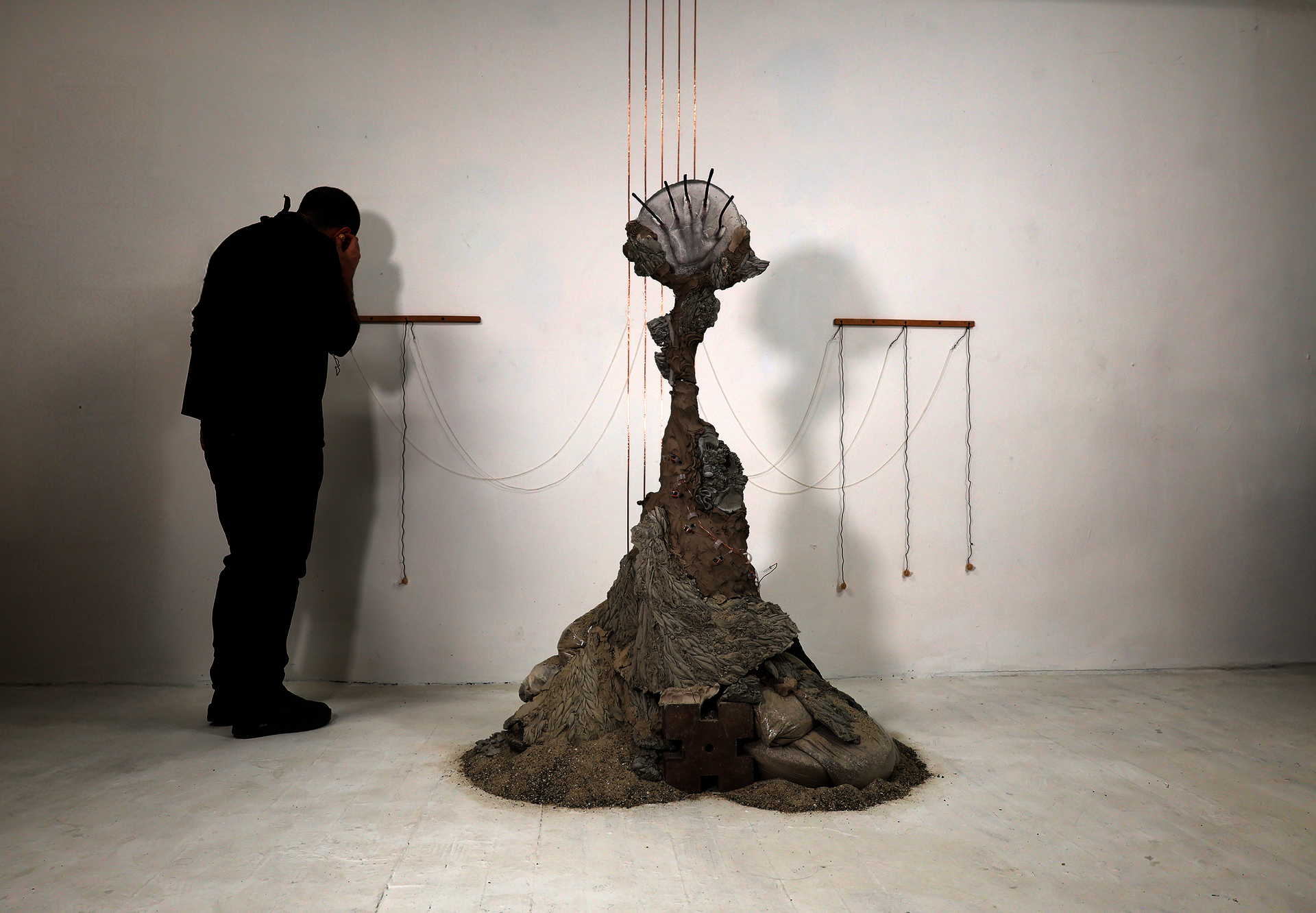 figure listening to mound of clay, concrete rubble, glass hand, copper wire around and up to the ceiling. sand on floor.