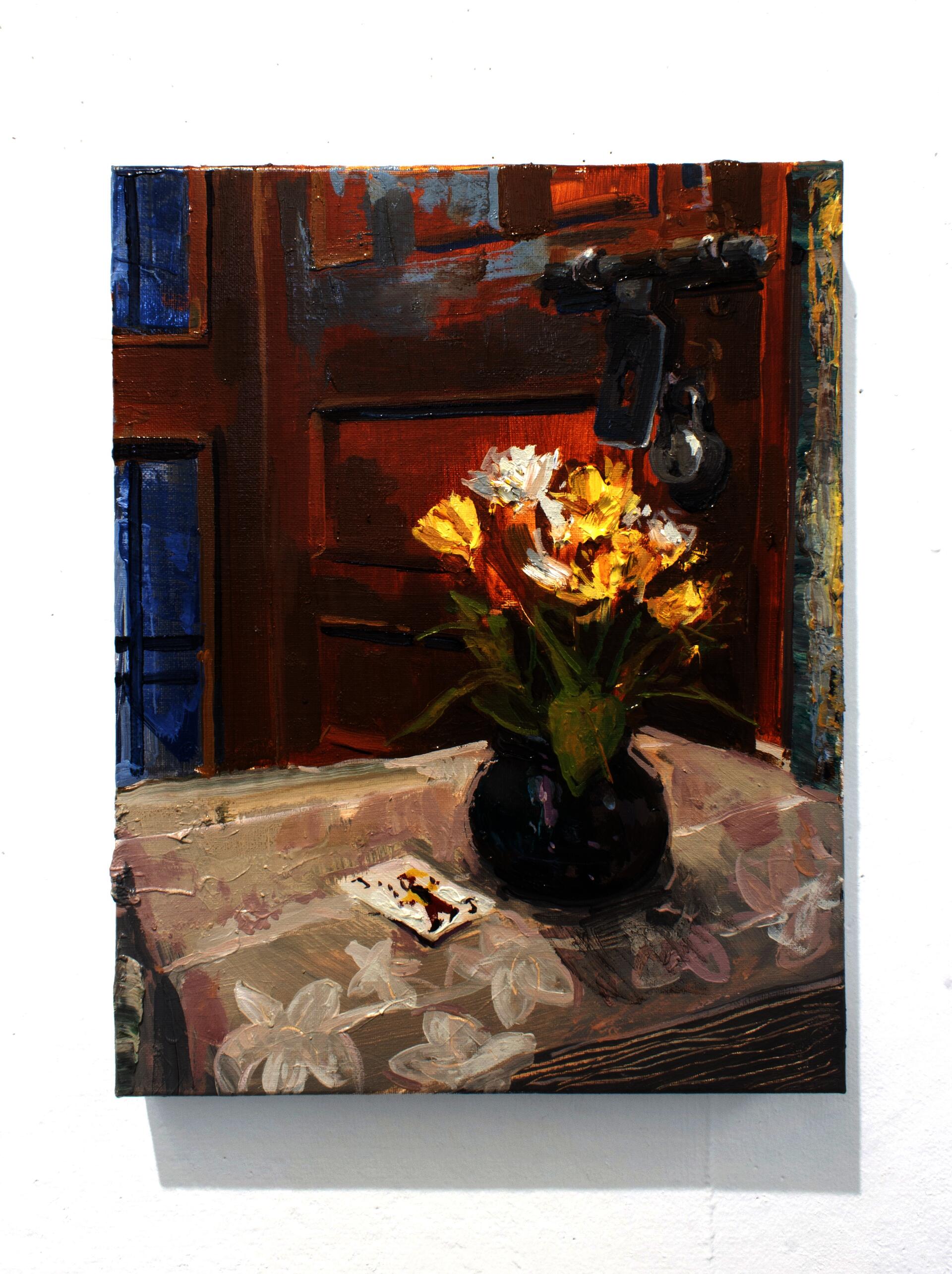 Yellow plastic flowers in a dark vase next to a joker card on a table with white floral tablecloth. Behind the vase is a wooden door with a lock.