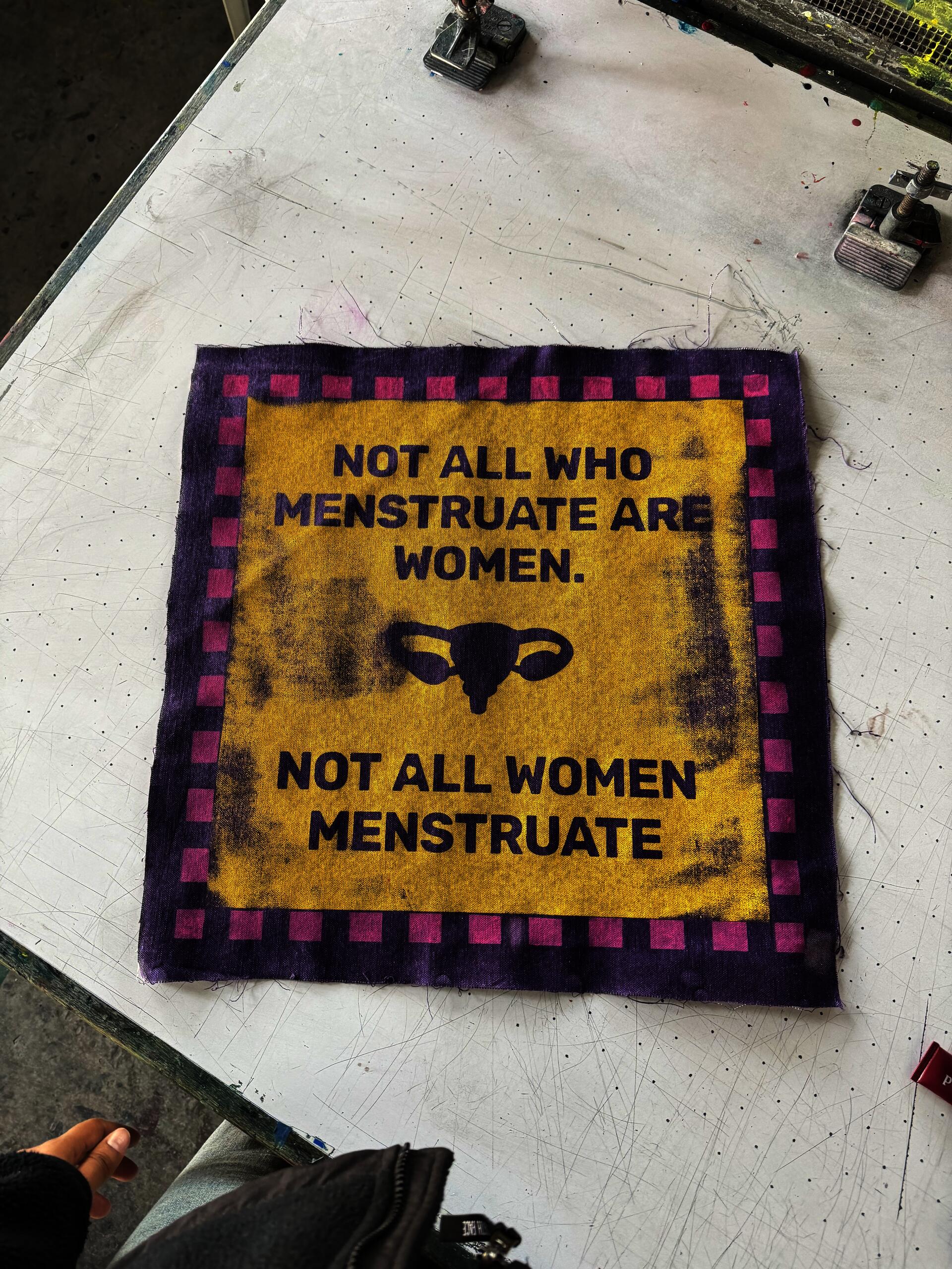 "Screen print on fabric, yellow, purple & pink A bold, graphic patch printed in purple on a golden-yellow field with a pink-and-purple checkered border. The text declares: ""Not All Who Menstruate Are Women. Not All Women Menstruate."" — flanked by a uterus icon. Photographed fresh off the print table."