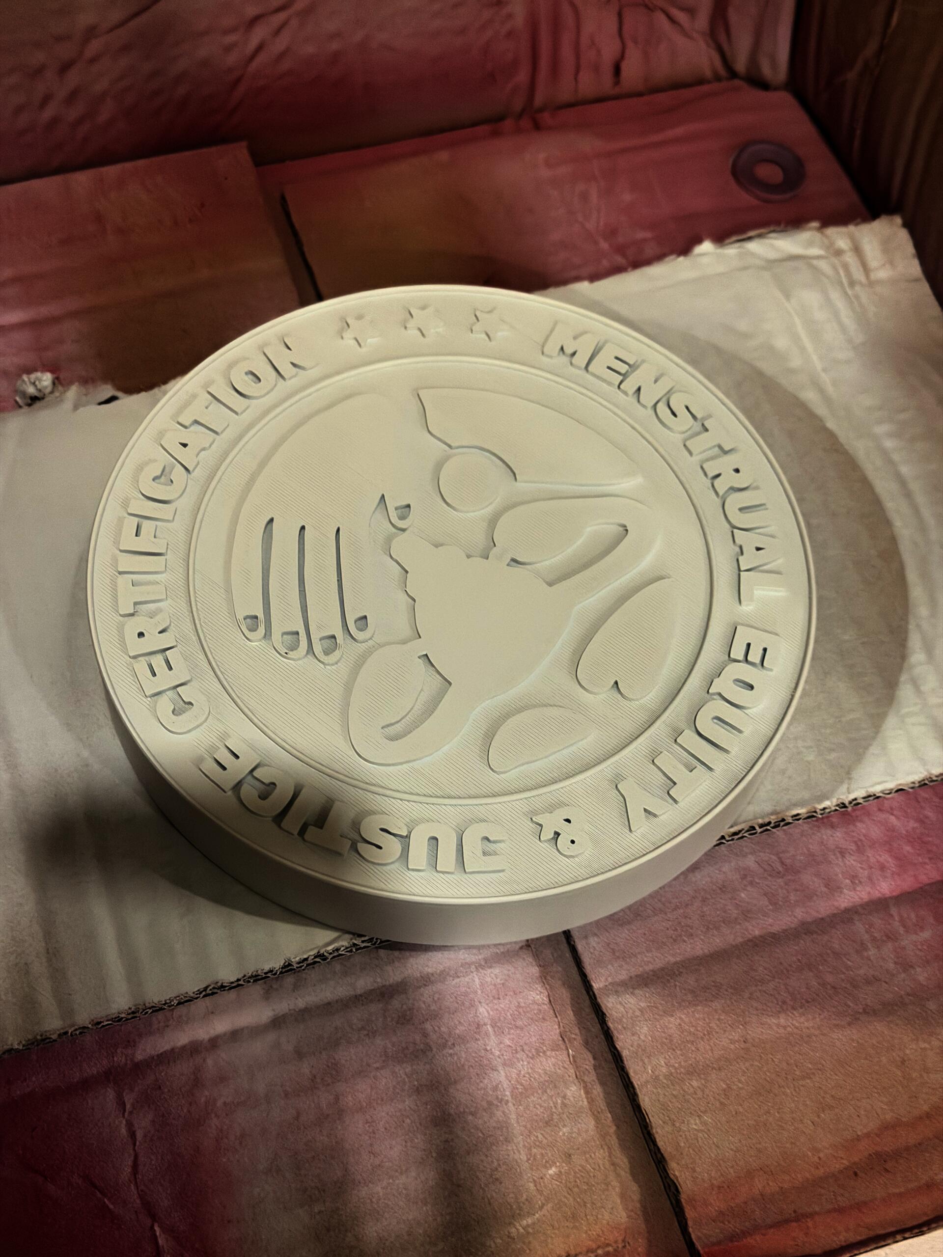 "3D print in white PLA A larger-scale version of the certification disc, resting on tissue paper and cardboard. The raised lettering and central imagery are clearly defined, showing the dimensional depth and layered relief of the 3D print."