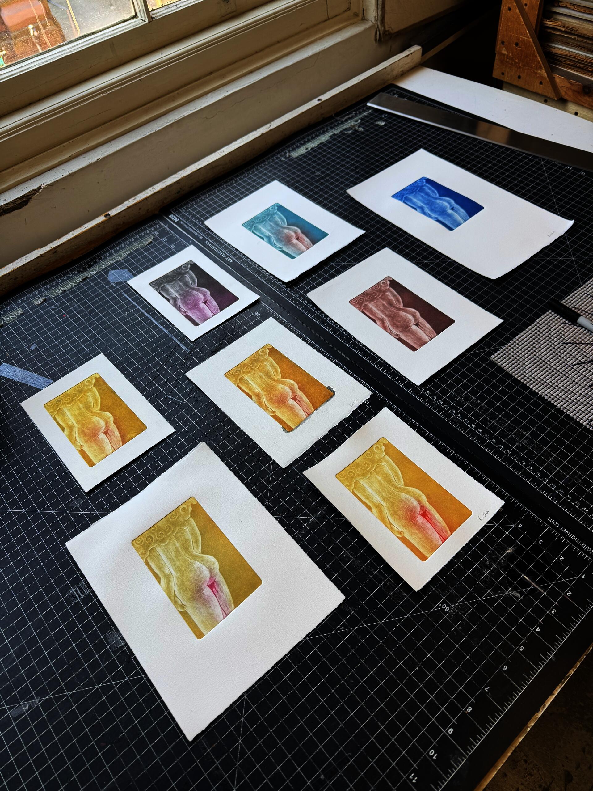 "Monoprints / relief prints on white paper, studio spread Seven prints laid out across a cutting mat, each featuring the same torso figure printed in a different colorway — teal, cobalt blue, mauve, warm brown, soft gold, and golden-rose. A studio documentation of an edition in progress, exploring how color shifts the emotional register of the same image."
