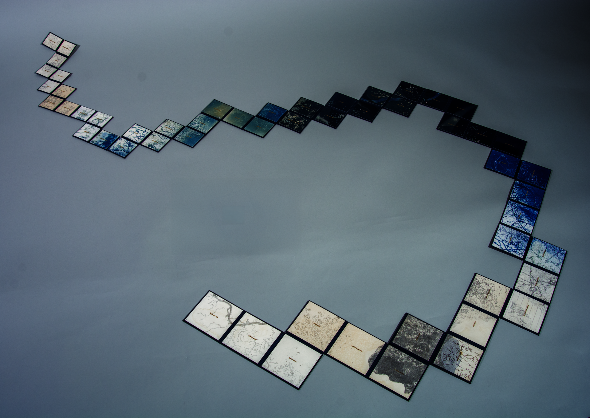 Squares of printed material ranging in color and tones of tan to dark blue are connected in a curved shape, receding in size