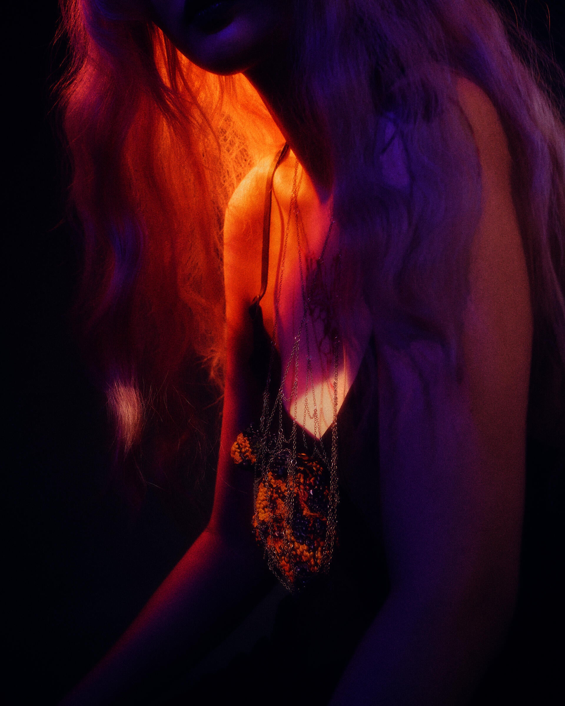 Close-up of a light-skinned figure in a black dress, angled left, wearing layered chains suspending a textured orange-brown heart-like pendant; saturated purple and warm amber lighting create an intimate, moody atmosphere.