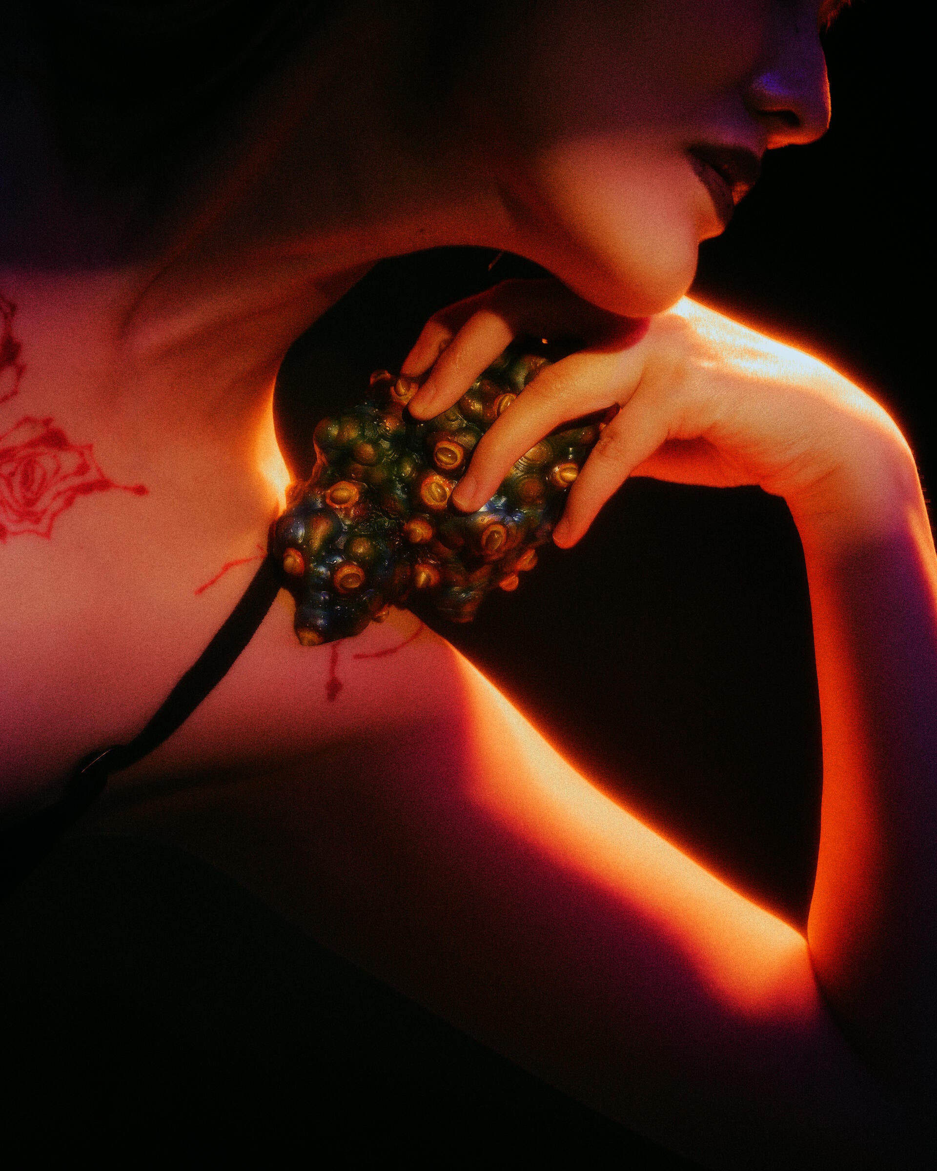Multicolored, textured heart-like object studded with raised cat’s eye stones rests on the shoulder, held by fingers; warm amber light highlights its surface while deep purple and green shadows create an intimate, tense mood.