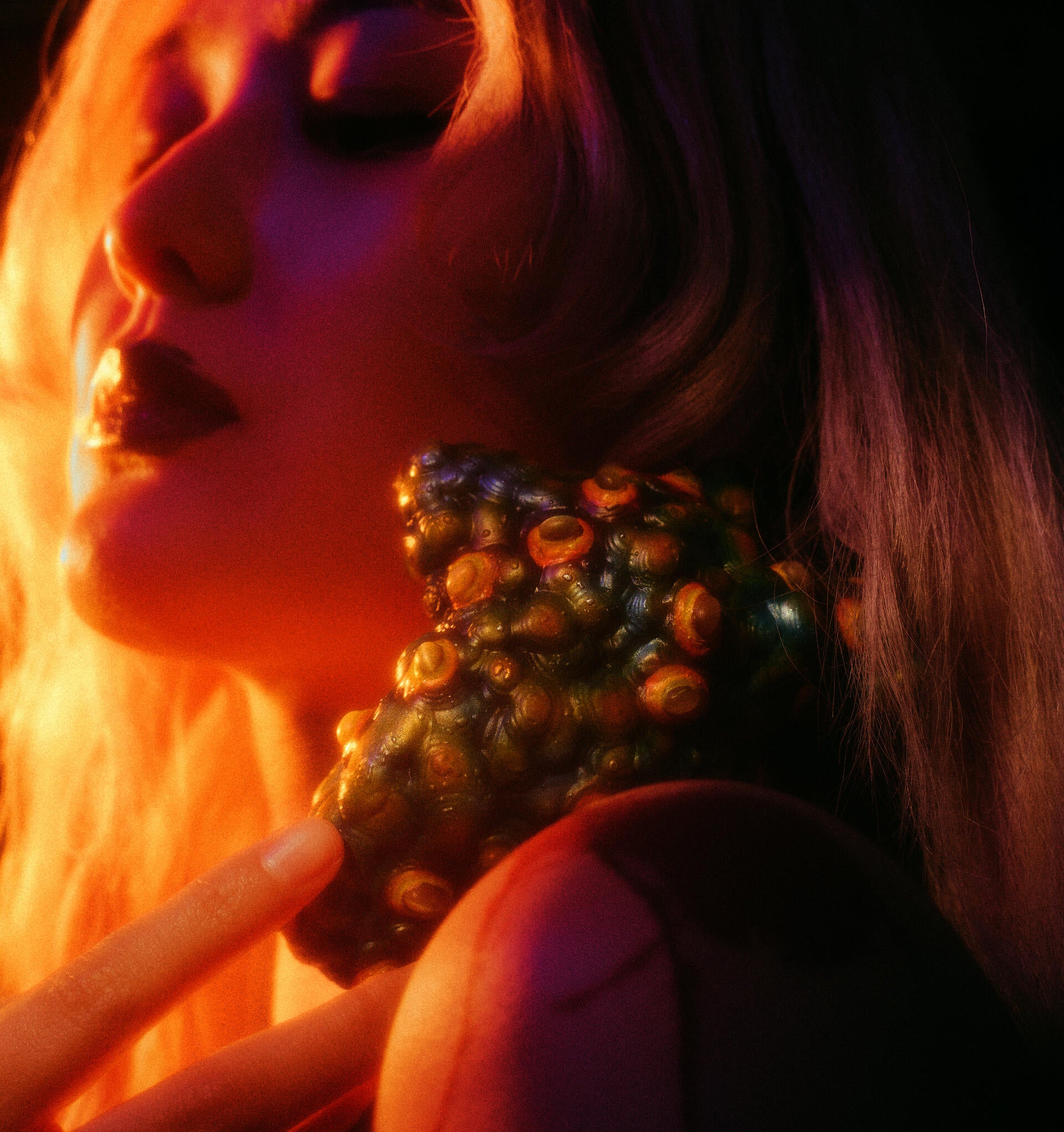Multicolored, textured heart-like object studded with raised cat’s eye stones rests at the neck and shoulder, touched by fingers; warm amber light highlights its surface while deep purple-green shadows create an intimate, tense mood.