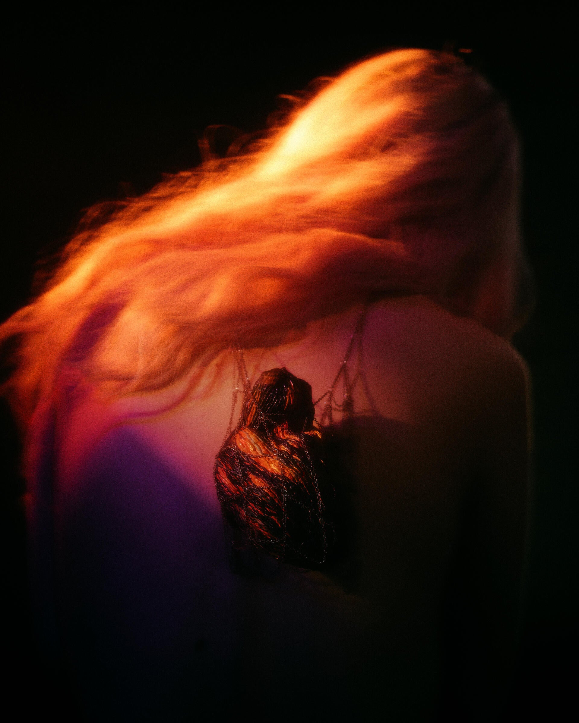 Heart-like pendant encased in fine chains hangs at center of the back, glowing amber-orange through a dark woven surface; cool purple shadows and soft green edges create a suspended, intimate tension.