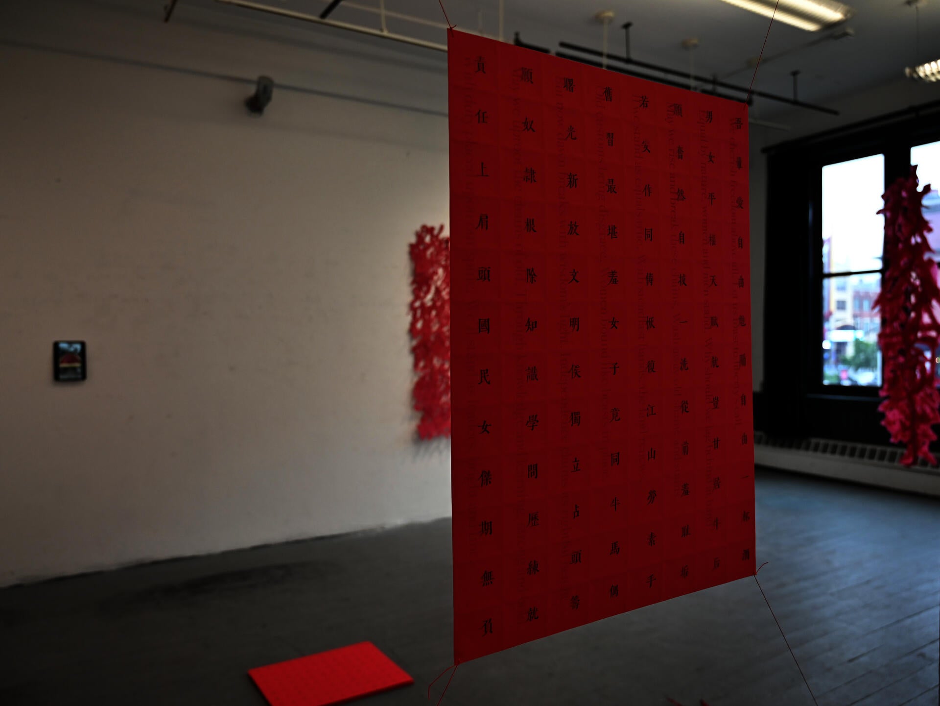 Red hanging paper sheet with vertical text, suspended in a gallery with red paper installations in the background.