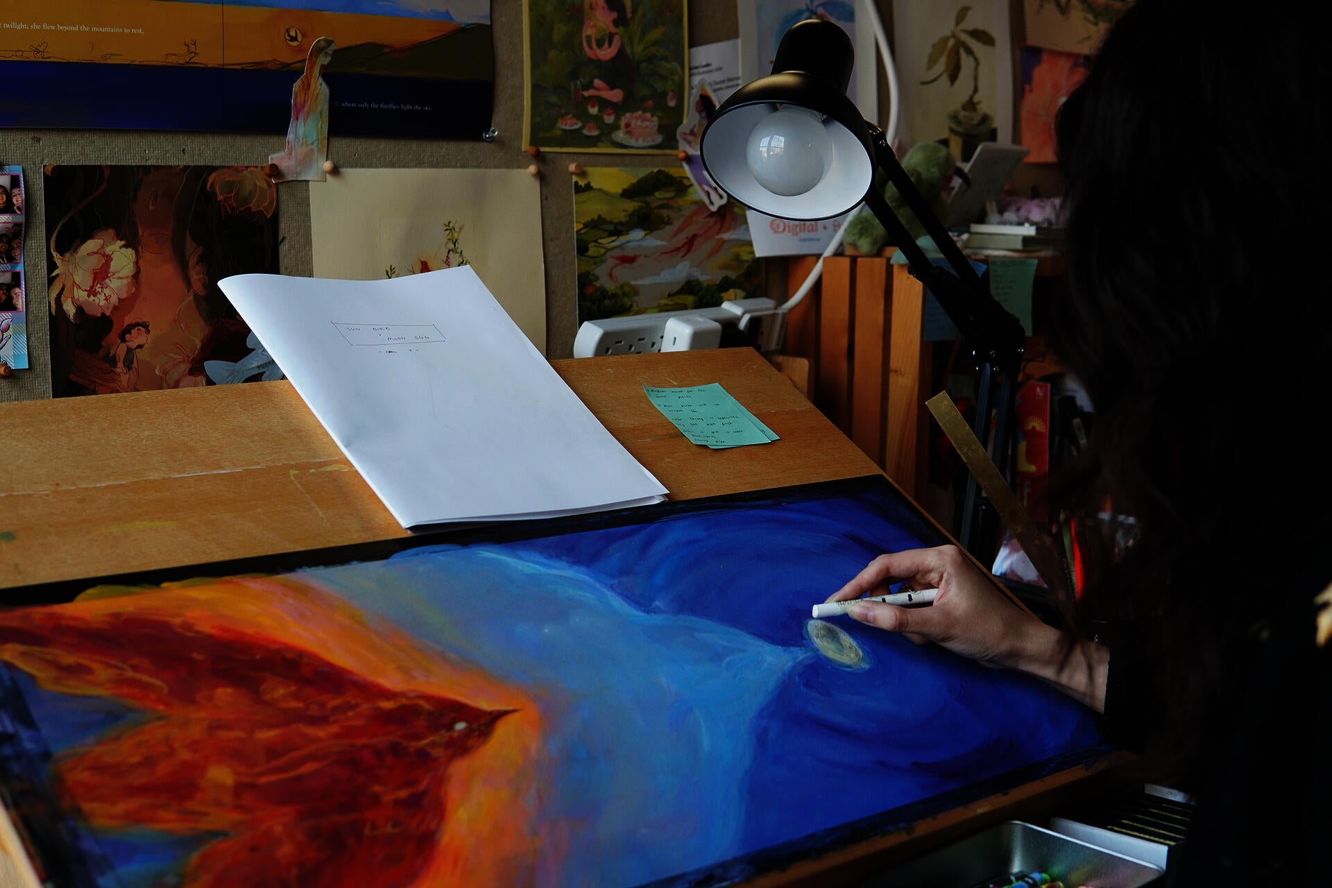 Photograph of the artist in her studio working on a picture book spread depicting a red bird flying from the left page toward a white egg against a dark blue night sky.