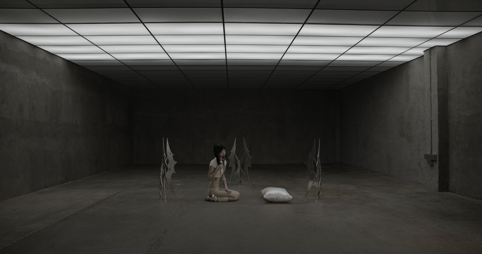 Wide shot of a person kneeling on concrete floor in a large gray room, surrounded by upright metal sculptures, holding hair near mouth; white pillow placed nearby under grid ceiling lights.