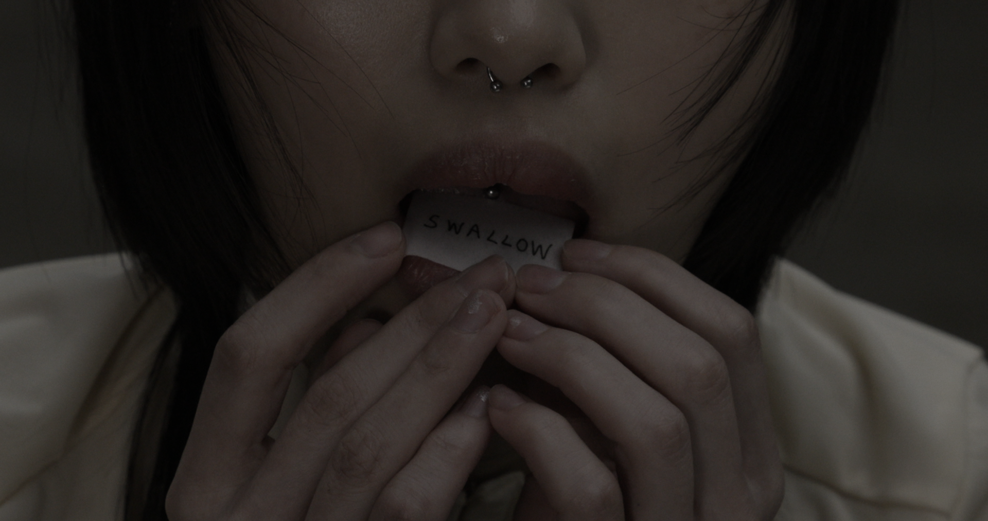 Close-up of mouth pressing a small paper labeled “SWALLOW” between lips; fingers hold edges of paper, skin and texture sharply visible.
