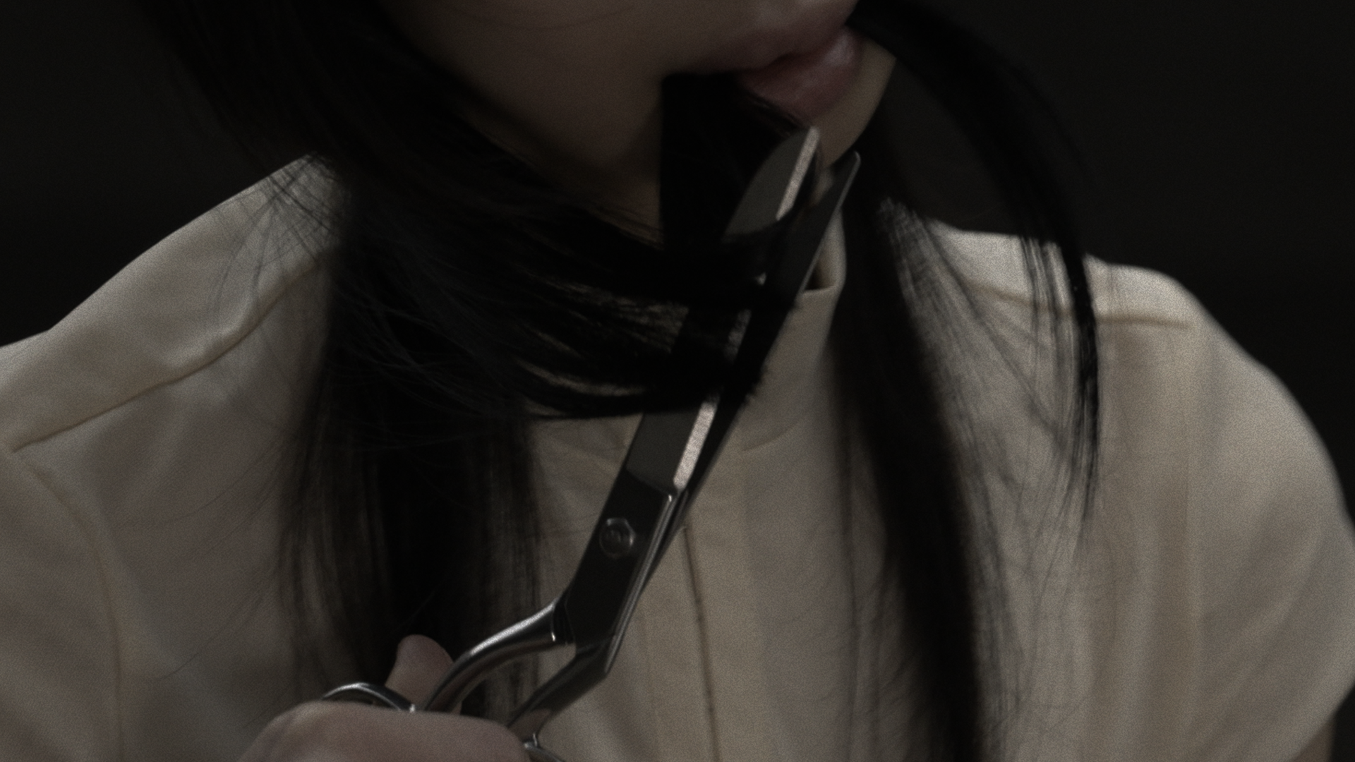 Close-up of scissors cutting a section of dark hair near the neck; strands are held taut between blades, with soft fabric clothing visible behind.